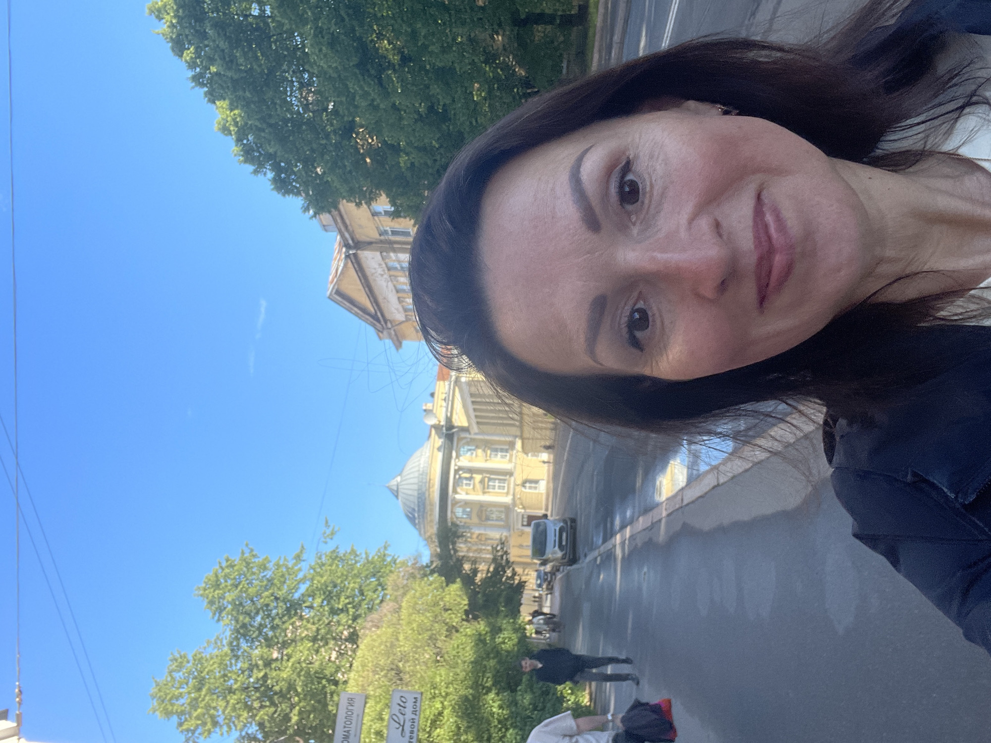 A woman is taking a selfie outdoors in Saint Petersburg, Russia. She is in the foreground, smiling. Behind her, a street scene unfolds. A sidewalk runs along the left side of the street, with trees lining it. People are walking on the sidewalk, and a car is visible on the street. A large yellow building with a dome is in the background. Signs in Russian are visible on the left side of the image. The sky is clear and blue.