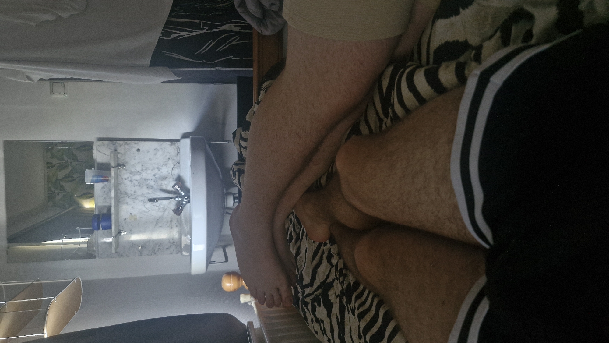 The image depicts a close-up view of a person's legs and lower torso, with a portion of a sink and mirror visible in the background. The person appears to be lying down, with their legs crossed and their body partially covered by a black garment with white stripes, possibly shorts. The background shows a white sink with a marble backsplash and a mirror reflecting a patterned shower curtain. The overall lighting suggests it might be daytime, though the exact time is unclear. The mood is relaxed and casual, with no strong emotions conveyed. No visible text is present in the image.