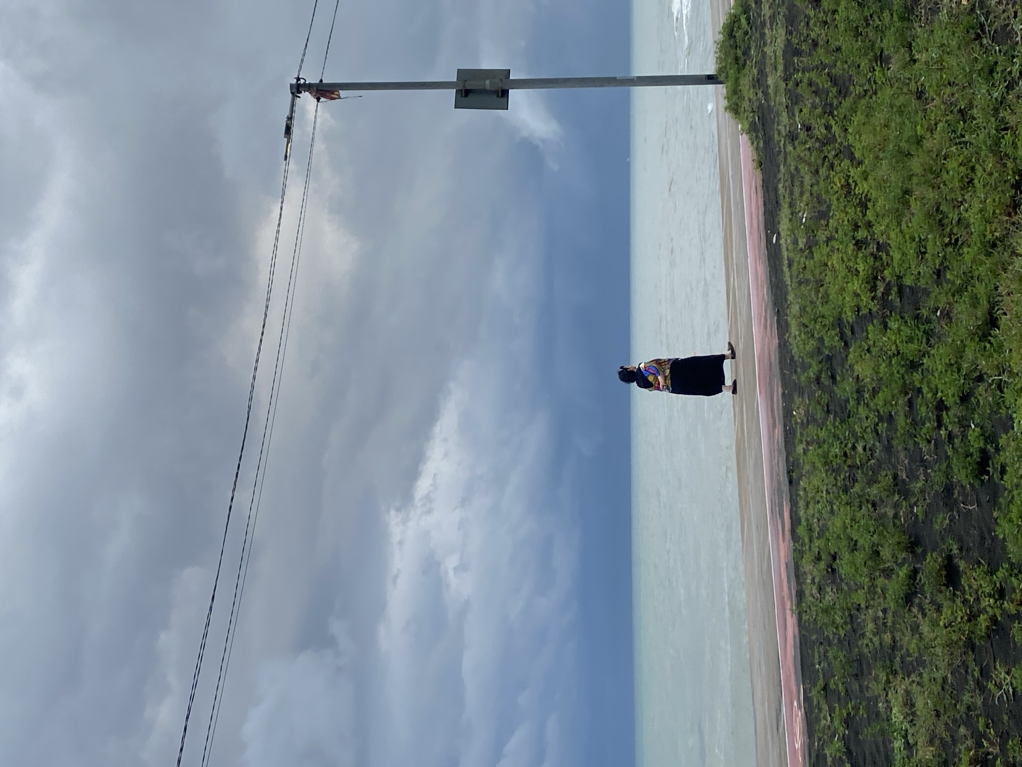 A single person walks along a promenade by the Black Sea in Ureki, Georgia. The sky is overcast with dark, dramatic clouds. The sea is a muted blue-green. The promenade has a red and white striped track. To the right, a utility pole stands with a sign attached and wires extending across the frame. Green grass and dark soil are visible in the foreground. The person, seen from the back, is wearing a long dark skirt and a colorful, patterned shawl or wrap.