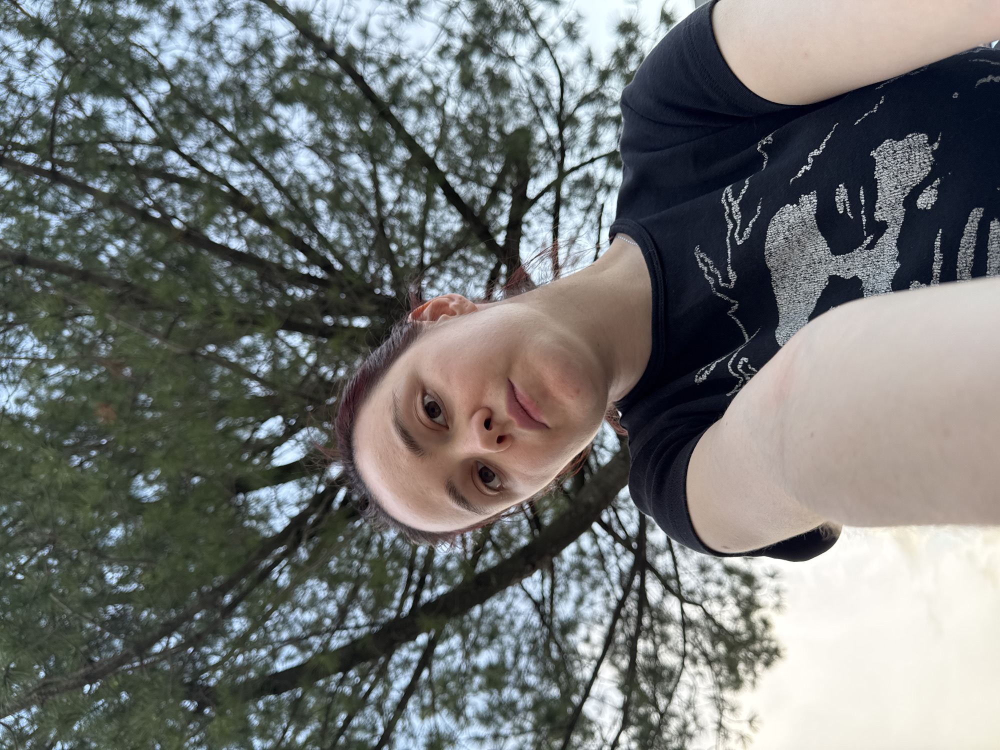 A selfie taken at a low angle shows a fair-skinned individual with auburn hair, wearing a black t-shirt with a pale graphic print. The person's face, with distinct eyebrows and brown eyes, is the primary subject. The background features a dense pine tree canopy against a light-colored, overcast sky. The location cannot be determined to be Izhevsk, Russia based on the information available in the image.