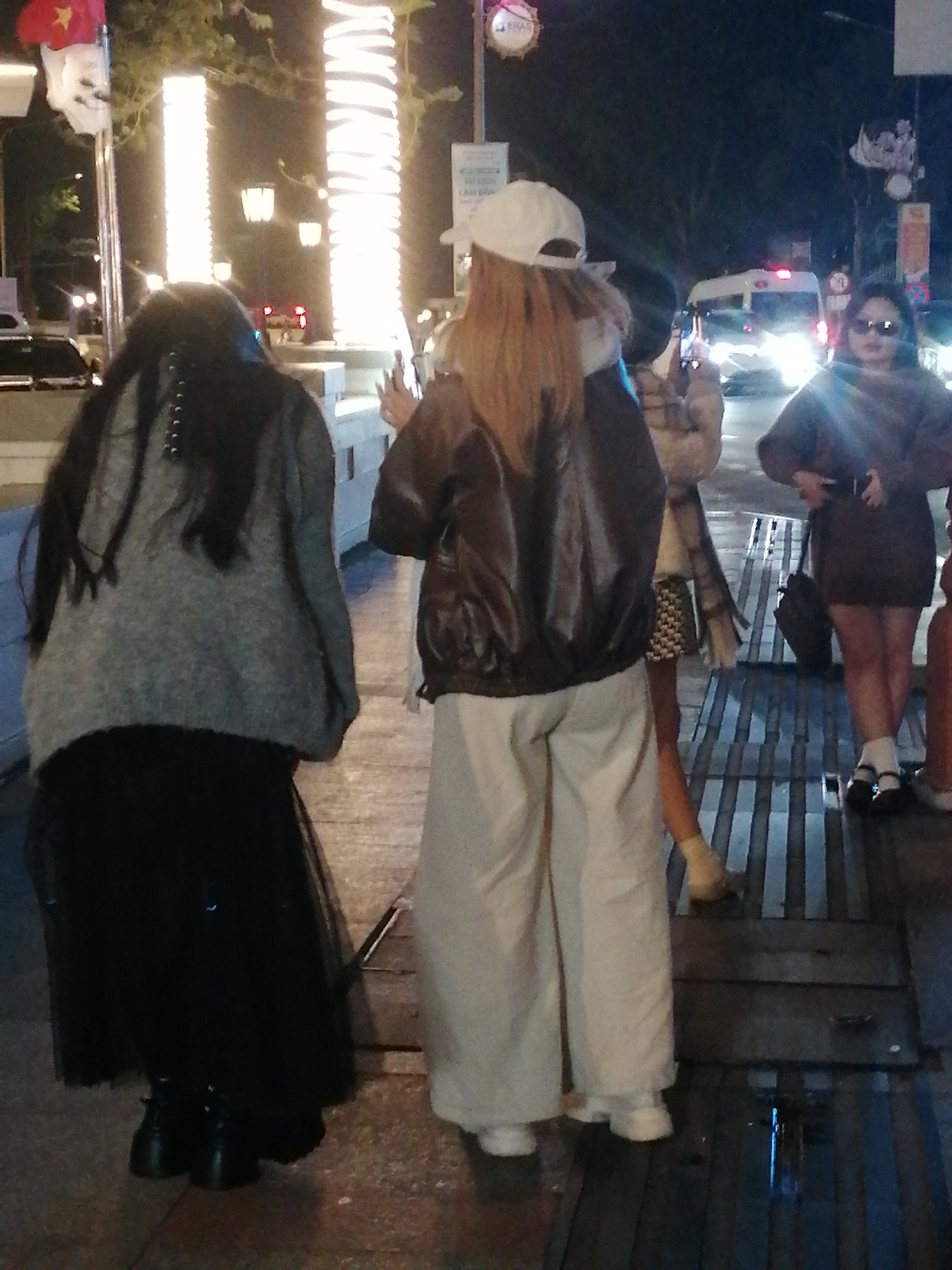 In Da Lat, Vietnam, at night, four individuals are visible outdoors on a street. The foreground features two individuals. The leftmost person, facing away from the camera, has long black hair and is wearing a grey sweater over a black skirt and black boots. To their right, a person facing away from the camera, wearing a white baseball cap, a dark bomber jacket, and light-colored wide-leg pants, is seen from the knees up. Behind them, a person in a light-colored jacket is partially visible. Further to the right, a fourth individual, also facing away from the camera, is wearing a dark brown sweater, sunglasses, and white socks with black shoes, with a dark bag on the ground nearby. In the background, a white van with its headlights on and red taillights is visible, along with streetlights and illuminated columns. The ground appears to be a paved walkway with metal grate elements.