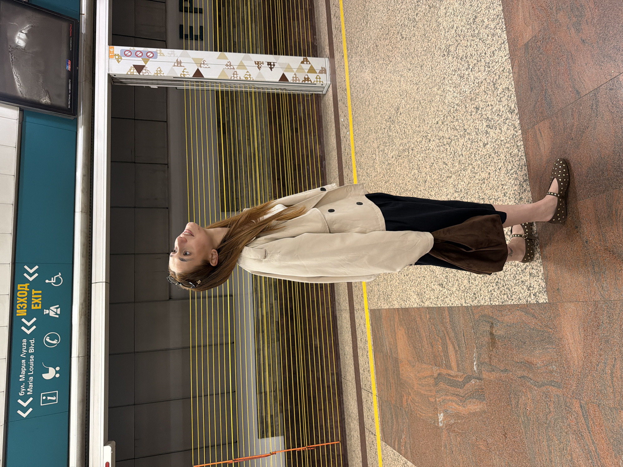The image depicts a woman standing in what appears to be a subway station in Sofia, Bulgaria. The woman has long brown hair, and is wearing sunglasses, a tan jacket, a black dress or skirt, and brown studded shoes. She is looking upwards and to her left.
The background includes a turquoise wall with the word "EXIT" in both English and Bulgarian, along with various symbols representing information, baby strollers, telephones, and accessibility. A sign indicates the location is "bul. Maria Luiza Blvd." There are yellow vertical lines, possibly a design element or a safety feature, along the wall. The floor is tiled with a mix of beige and red-brown stone tiles. A display screen and decorative geometric patterns are also visible in the background.