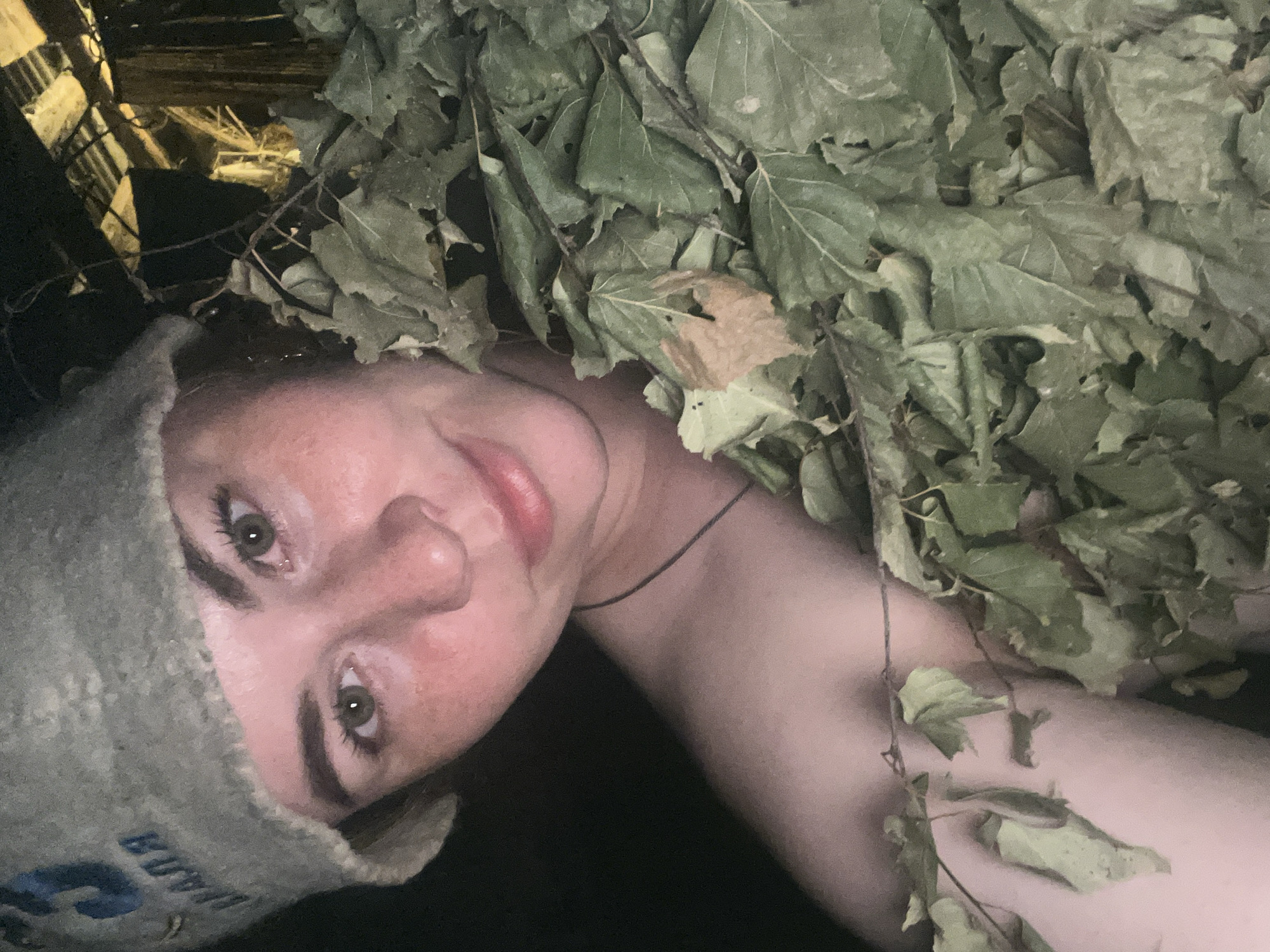 A person with fair skin is positioned at an angle in a dark environment. They wear a gray felt hat with Cyrillic lettering on the left side of their head. The person is surrounded by green leaves, obscuring much of their body. A thin, dark string is visible around their neck. In the upper left corner, there are glimpses of wooden planks illuminated by a light source. This photo may be from Сосново, Russia.