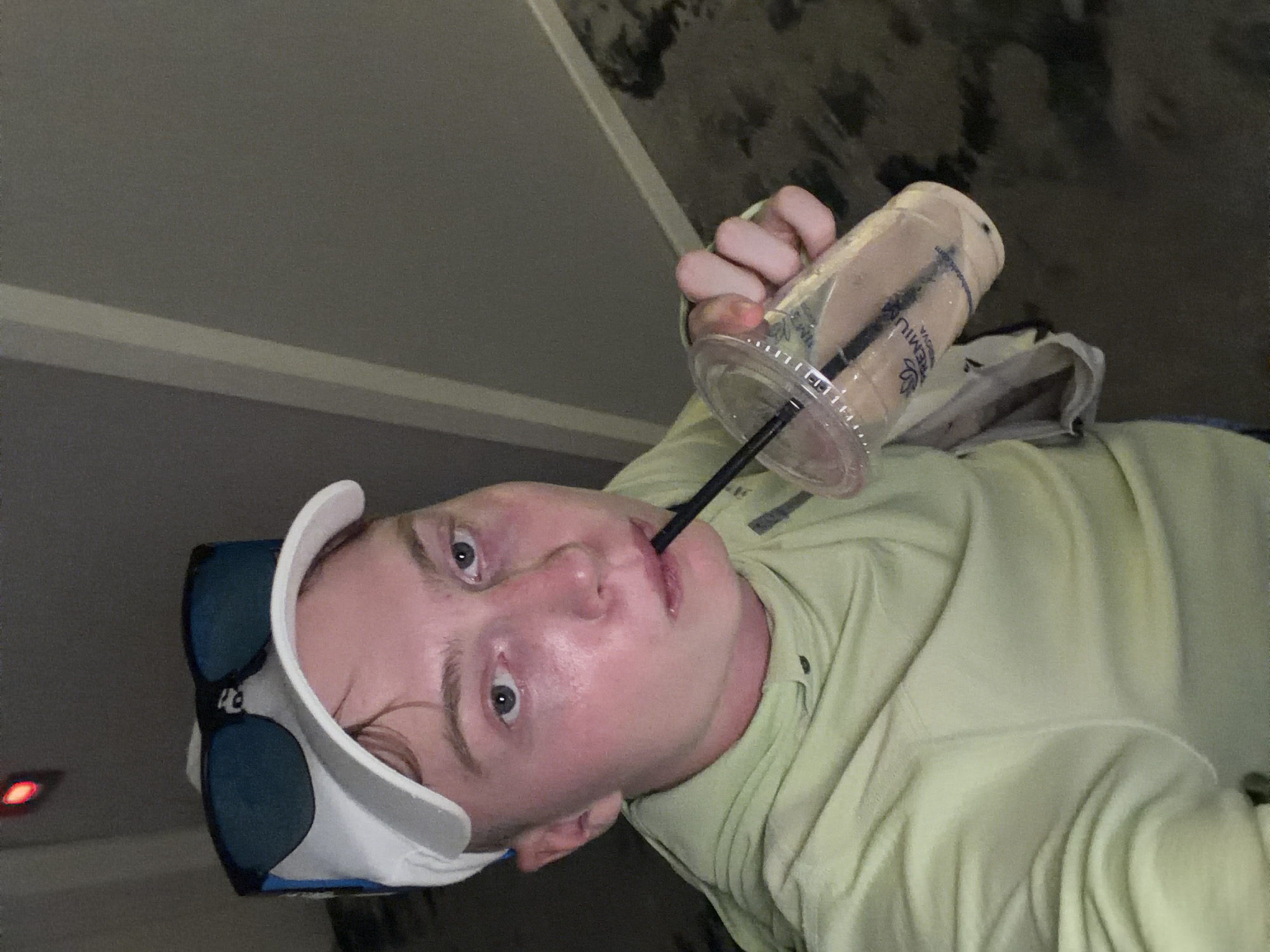 A person is shown from the chest up, indoors, holding a clear plastic cup with a dark liquid and a black straw to their mouth. They are wearing a light green hooded sweatshirt, a white baseball cap, and blue-tinted sunglasses perched on top of the cap. The background appears to be a hallway with grey walls and a patterned floor. The person's face is flushed, and their eyes are open, looking towards the camera. The lighting is dim.