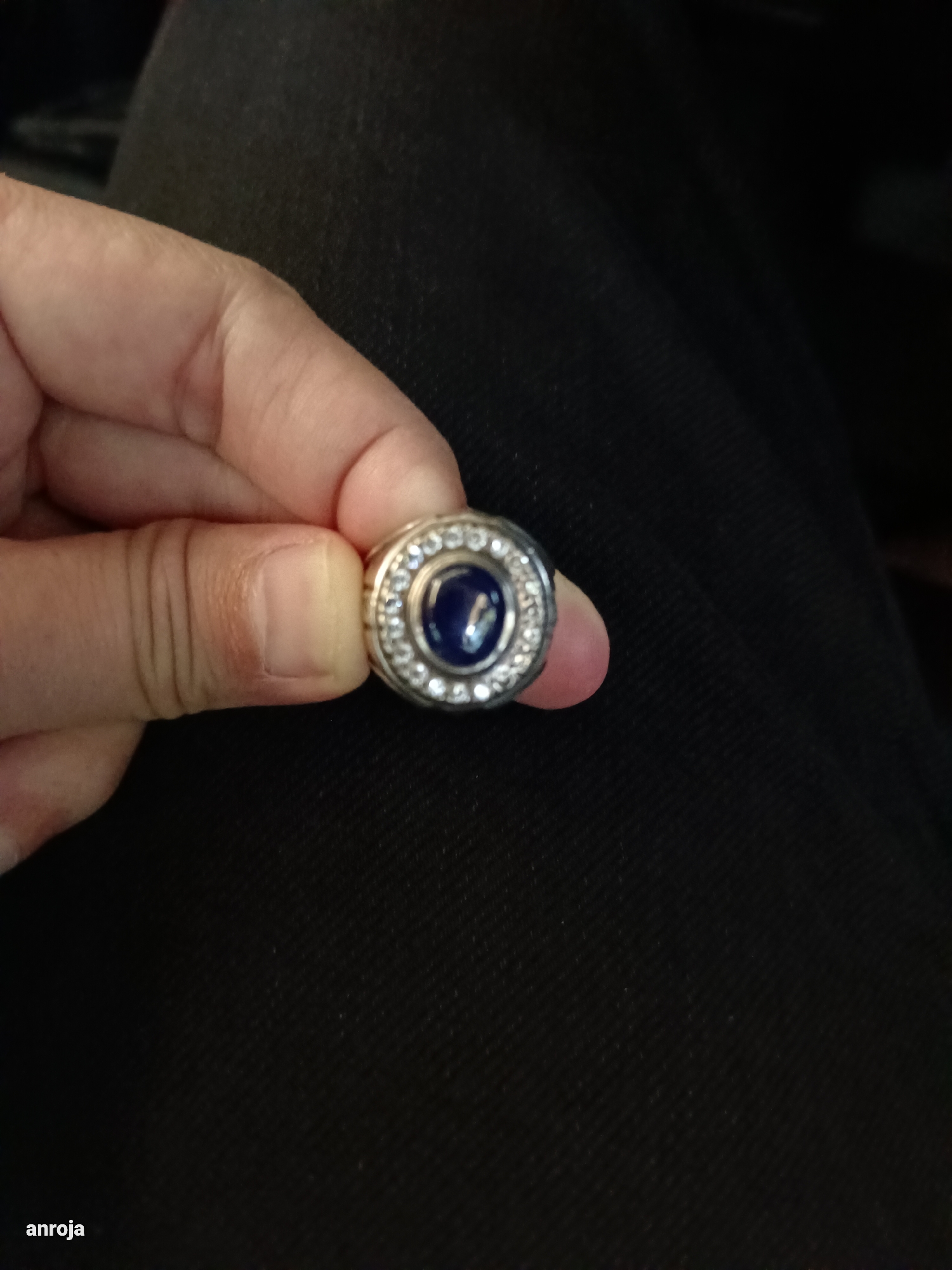 An individual's left hand is holding a silver ring with a blue gemstone in the center, surrounded by small clear stones. The ring is positioned on a dark, textured surface, likely fabric. The background is dark and out of focus. The image is cropped from a larger scene, and the location in Blang Cut, Indonesia is not discernible from the visual information provided. The watermark "anroja" is visible in the bottom left corner.