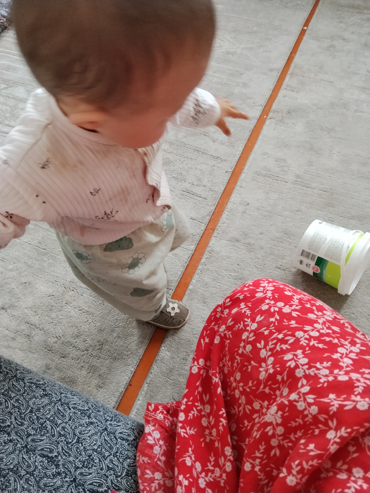 The image shows a close-up, overhead view of a baby in a light-colored, long-sleeved shirt and light-colored pants with a floral pattern. The baby is wearing a single gray shoe with a white soccer ball design. The baby's head is turned away from the camera, and one hand is extended forward. To the right, a white plastic container with green and yellow labeling is lying on its side on a gray carpet. In the foreground and right side of the image, a person wearing a red floral-patterned skirt or dress is partially visible. To the left and foreground, a portion of a dark blue fabric with a white paisley pattern is visible. The carpet has an orange strip running diagonally across it. The scene appears to be indoors.