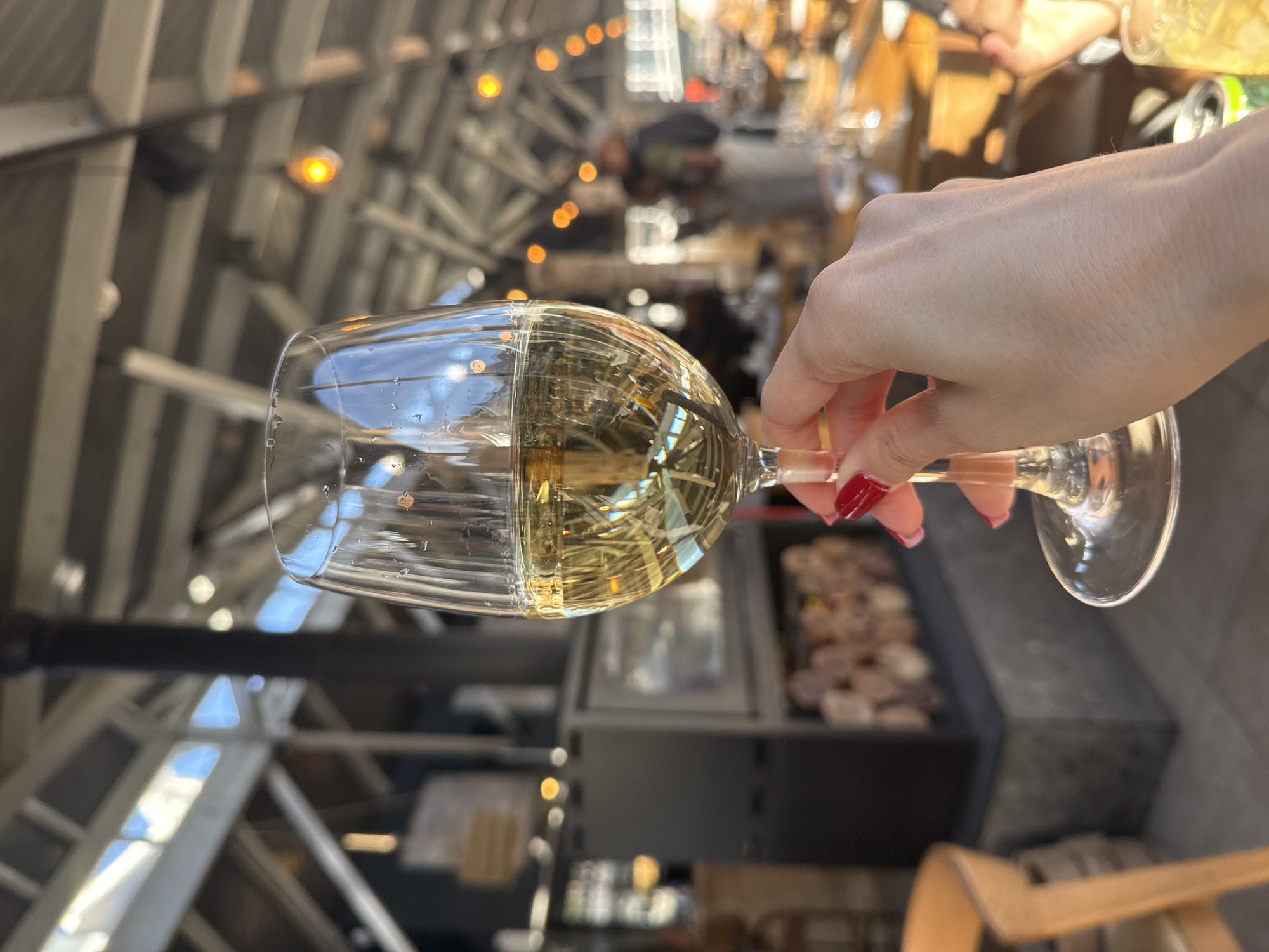 A person's hand, adorned with bright red nail polish, holds a clear wine glass filled with a pale golden or light-yellow liquid, likely white wine or sparkling wine. The glass is positioned in the foreground, with reflections on its surface revealing parts of the background structure.
The setting appears to be a spacious, modern indoor or semi-covered area with an industrial aesthetic. The background features a high ceiling with an intricate pattern of exposed light-colored beams and girders, indicative of a renovated warehouse or a contemporary architectural design. Numerous warm-toned, small incandescent lights are strung across the ceiling, contributing to a lively and inviting atmosphere. Interspersed among the structural elements are areas that suggest natural light coming from windows or skylights.
In the mid-ground, behind the glass, blurred elements hint at shelves or display cases, possibly showcasing food items or products, consistent with a market, food hall, or a large bar/restaurant environment. The activity depicted is simply enjoying a drink, suggesting a leisurely moment in a social or public setting. The overall mood is relaxed and pleasant.
Considering the location context of Cape Town, this scene could be from one of the city's popular food markets, wineries, or bustling eateries known for their modern design and vibrant social scene. No discernible text is visible in the image.
