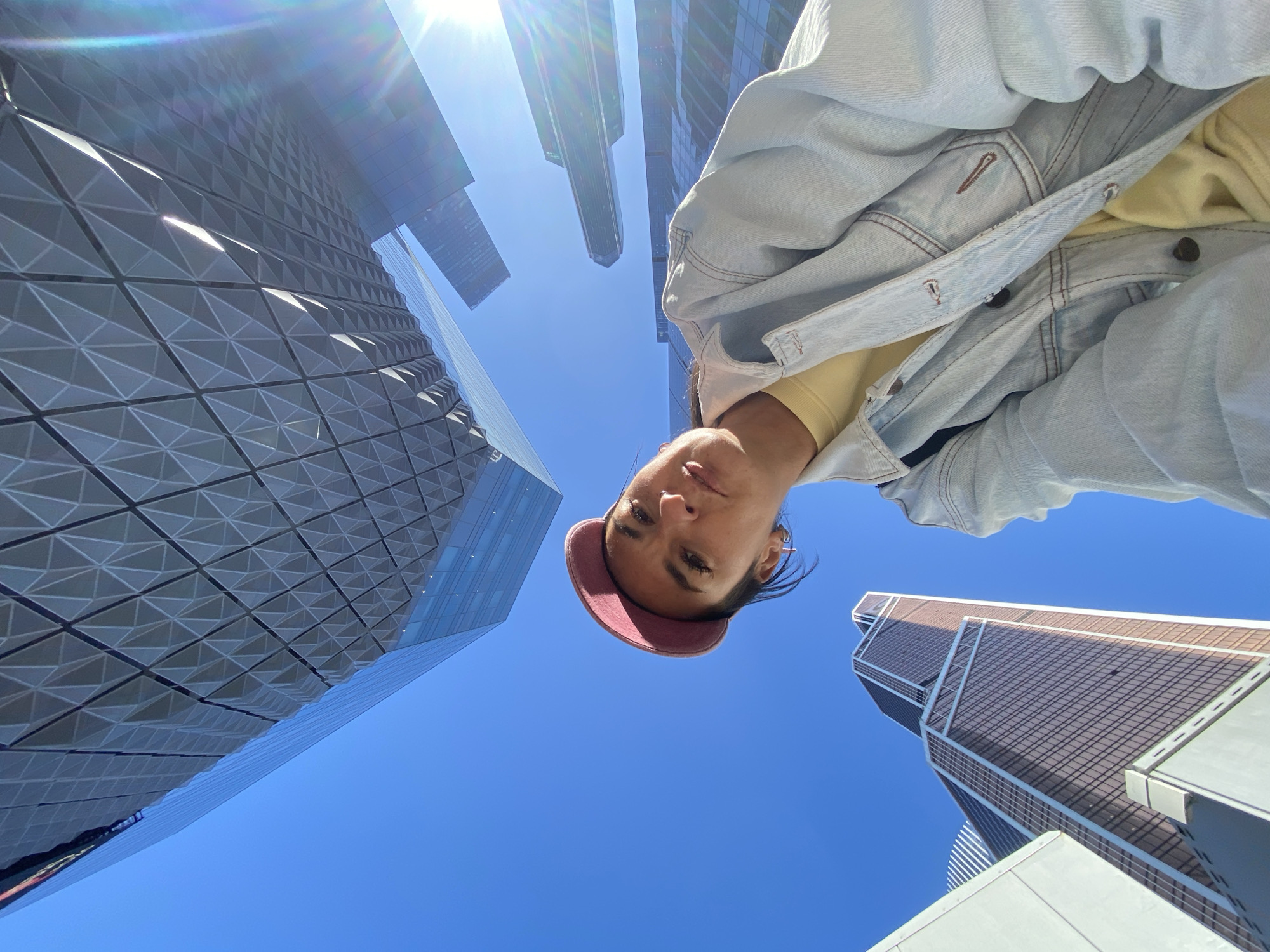 A low-angle shot features a woman in a light blue denim jacket and a red baseball cap, looking down at the camera. Above and around her are several modern skyscrapers, partially visible. The sky is a clear, bright blue, and the sun is visible in the upper part of the frame. This view is consistent with the location in the city of Khimki, Russia.