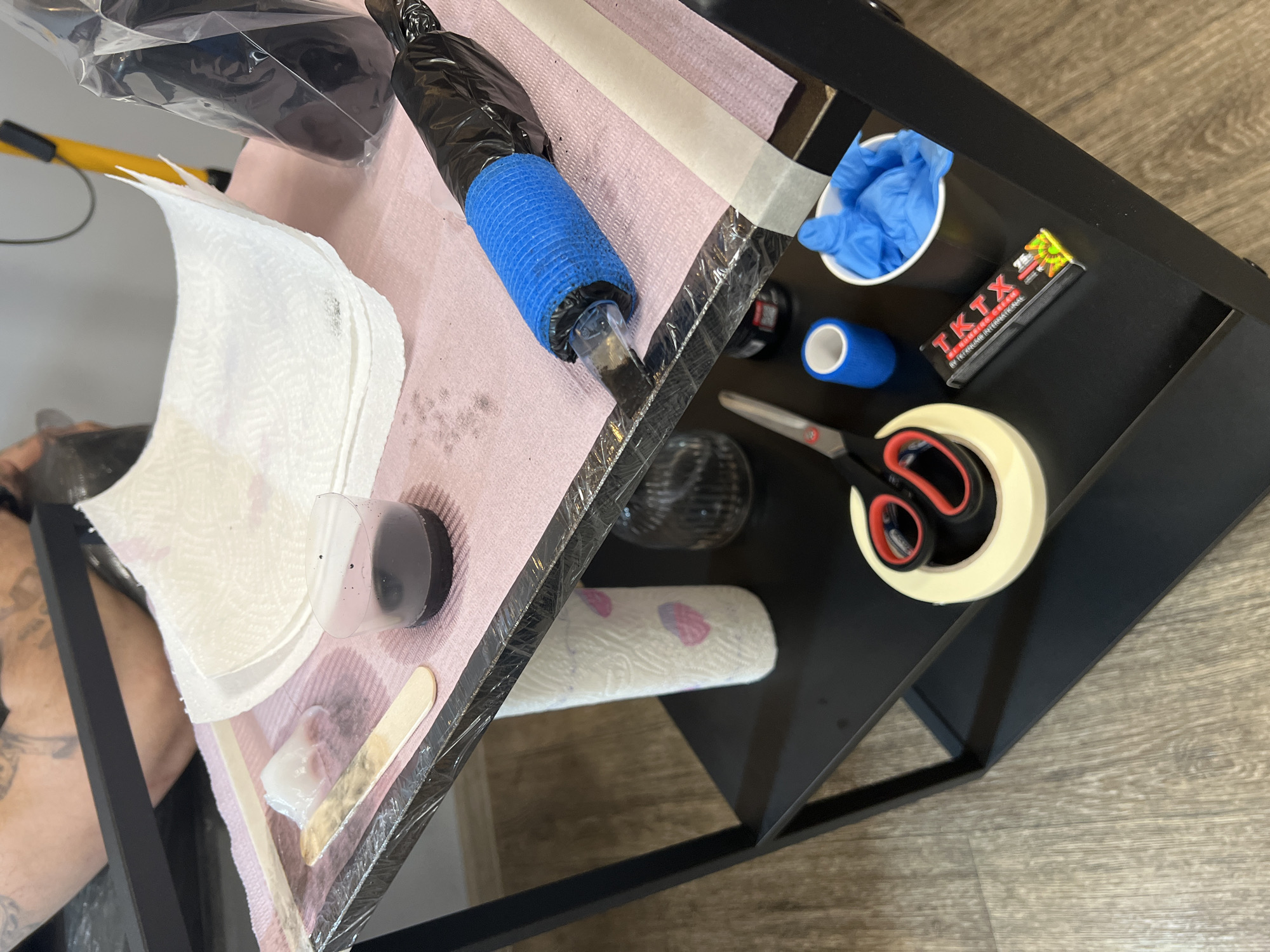 This media file captures a detailed, overhead view of a tattoo artist's workstation, likely located in a studio in Saint Petersburg, Russia. The scene focuses on the tools and supplies necessary for a tattooing session, arranged on a black multi-tiered utility cart.
The main subject is the setup itself, with a glimpse of a person's arm (possibly the client's or artist's) on the far left, showing existing tattoos.
Key objects include a tattoo machine, centrally positioned, wrapped in black plastic and blue self-adhesive grip tape. It is placed on a pink disposable barrier, which also holds several ink caps (one containing dark ink), a stack of white paper towels, a wooden mixing stick, and a dab of ointment. On the lower shelf of the cart, there's a white cup filled with blue nitrile gloves, a roll of blue self-adhesive bandage, a small black box labeled "TKTX," a roll of white medical tape, and a pair of scissors with red handles. Another roll of paper towels is also visible on the lower shelf.
The activity depicted is the preparation for or the actual process of tattooing. The organized and sterile appearance of the workstation, with disposable barriers and ready-to-use equipment, suggests a professional tattoo environment.
Notable details include the clear emphasis on hygiene through the use of plastic wraps and disposable surfaces. The lighting is typical of an indoor setting, with no specific time of day or weather cues. The visible text on the black box clearly reads "TKTX" along with "DEEP NUMBING INTAGEN INTERNATIONAL," indicating a numbing cream product. The overall impression is one of readiness and professional execution within a tattoo studio.