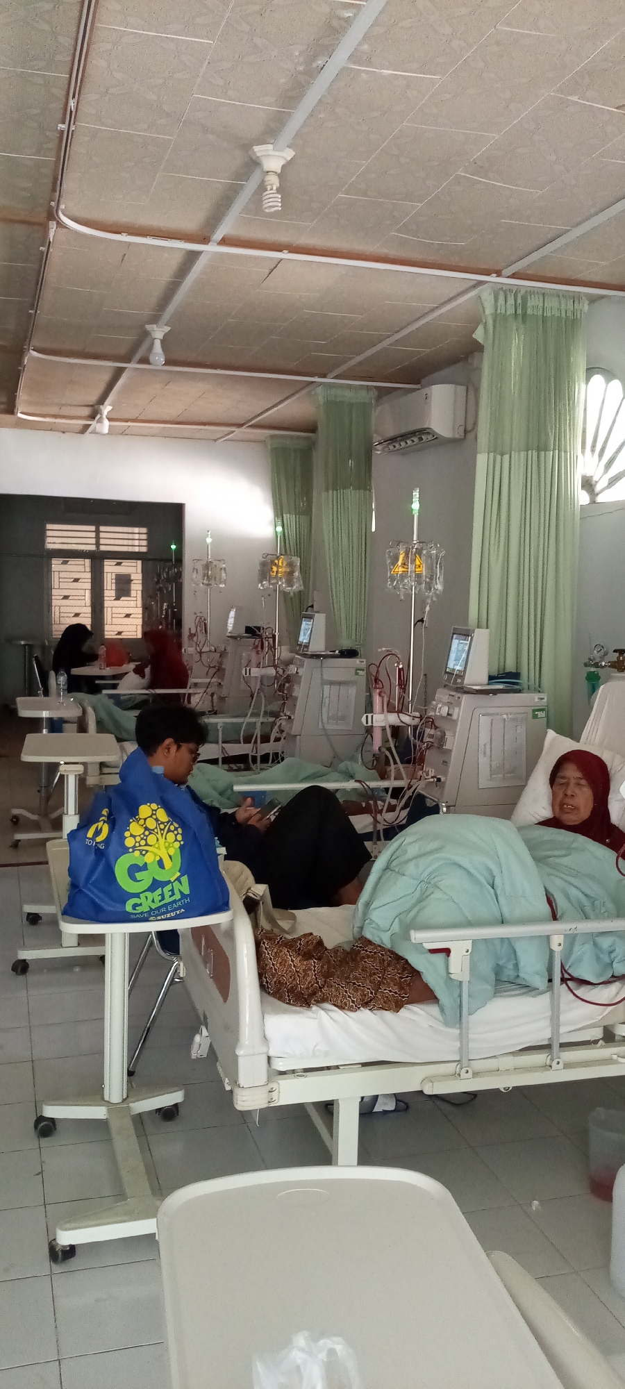 This image depicts a hospital room in Lhokseumawe, Indonesia, likely a dialysis center, where several patients are undergoing treatment. The room is equipped with multiple dialysis machines, each with accompanying IV stands holding bags of fluid. Green curtains are drawn, separating different patient areas. The ceiling is tiled and features fluorescent lighting fixtures and an air conditioning unit.
The primary subjects are several individuals in hospital beds. In the foreground, a young person with short dark hair is seated on the edge of a bed, holding a smartphone and looking at it. They are wearing a dark hoodie and a bright blue bag with yellow lettering is placed on a nearby stand. To their right, a patient is lying in a bed, covered with a light blue blanket and a patterned brown blanket. Another patient, wearing a red hijab, is also lying in a bed, her eyes appear closed. In the background, other patients and medical equipment are visible.
The activity taking place is clearly dialysis treatment, indicated by the presence of the dialysis machines and IV drips connected to the patients. The atmosphere appears calm, with patients resting or engaged with their phones. The lighting suggests it is daytime, though the closed curtains prevent clear observation of outdoor weather conditions. The location cues, such as the style of the beds, medical equipment, and the air conditioning unit, are consistent with a healthcare facility in Indonesia. No prominent text is discernible other than on the blue bag.
