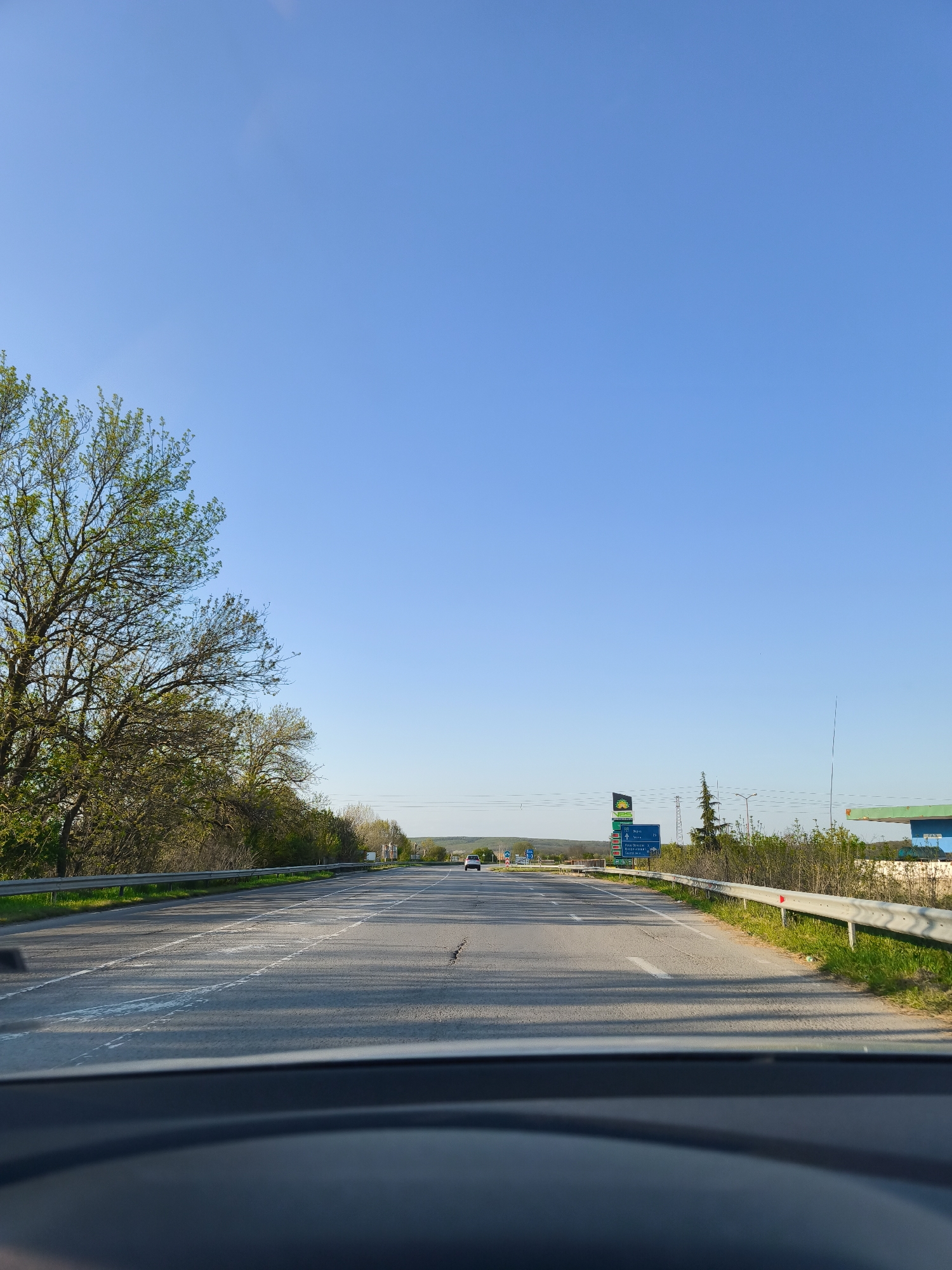 A view from inside a car looking out onto a long, straight highway. The road is lined with trees and guardrails on both sides. A sign with multiple Bulgarian city names is visible in the distance on the right side of the road. The sky is clear and blue. The car's dashboard is visible at the bottom of the frame. Based on the presence of road signs in Bulgarian and the request to provide a location, the image is likely taken on a road somewhere nearby Staro Oriahovo, Bulgaria. The scene is a view of the road, with no people visible.
