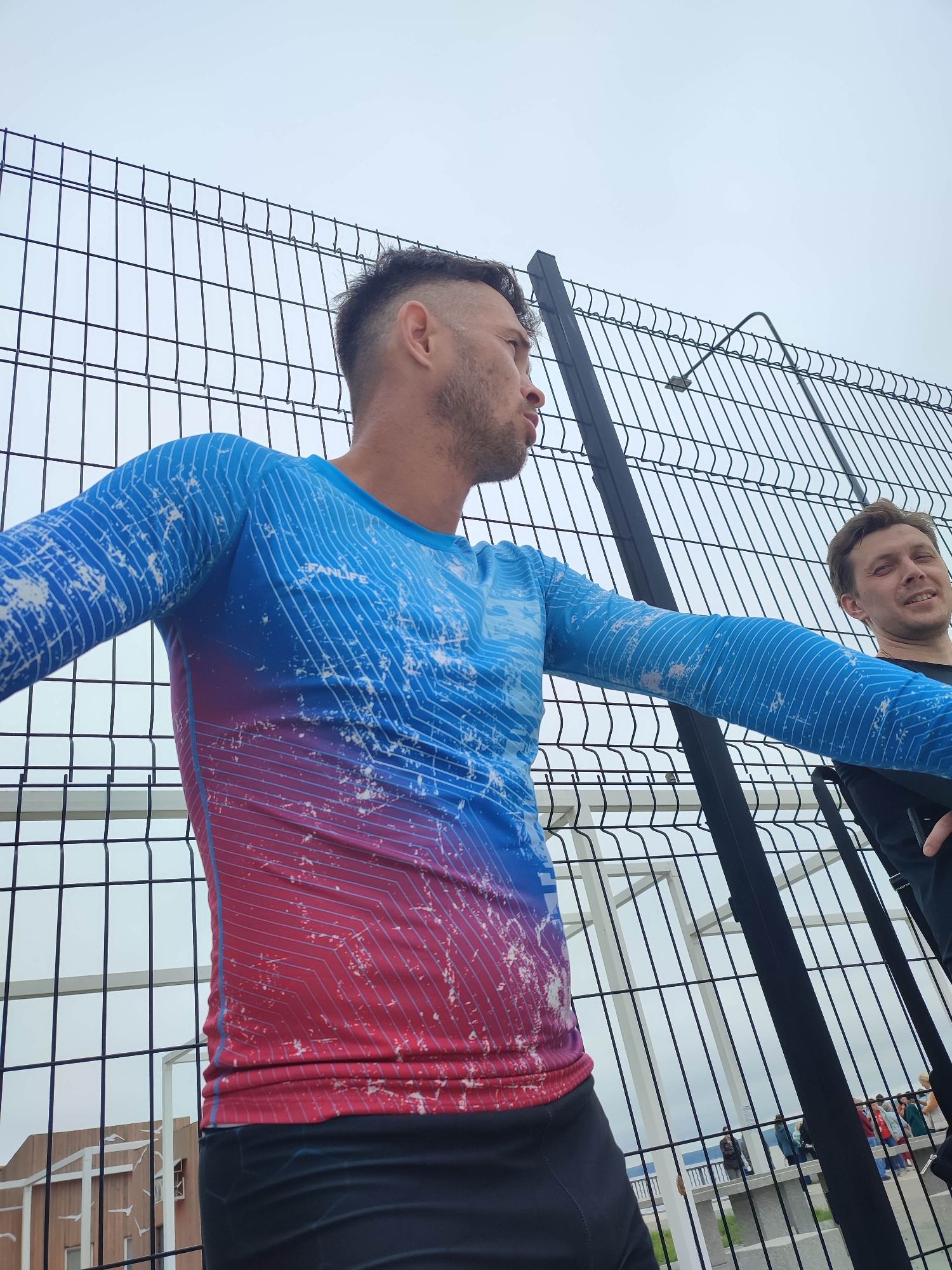 Two men are standing near a fence. The man in the foreground is wearing a long-sleeved blue and red shirt with a white line pattern. He has short dark hair and is looking to the right. The man in the background is wearing a black shirt and is smiling and looking to the left. The fence is black and has vertical bars. The sky is overcast.