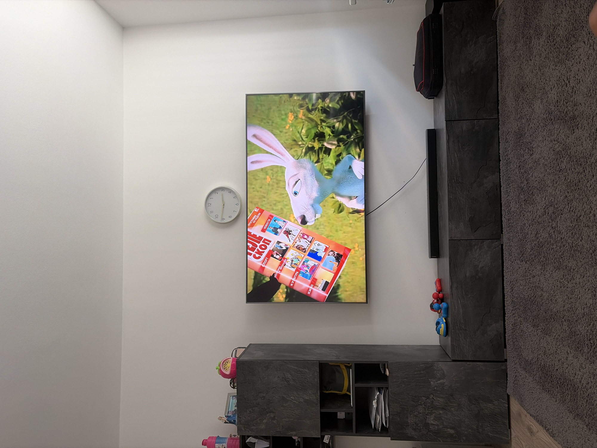 A television screen displays a cartoon image of a white rabbit with large ears in front of a green, leafy background. A red and orange magazine, with the title "ТЕЛЕ СКОП" visible, is being held up in front of the screen, partially obscuring it. The magazine cover features a grid of smaller images depicting various scenes, some appearing to be cartoons or illustrations. Above the television, a circular wall clock with a white face and black hands is mounted on a white wall. To the left of the television is a tall, dark gray cabinet with multiple shelves and drawers, containing various items including papers and what appears to be a bag. To the right of the television, on a dark gray media console, are a few children's toys, including a blue shoe, a red toy car, and a yellow and blue toy. A black soundbar is positioned in front of the media console, and a black case with red trim rests on the far right side of the console. The floor is covered with a dark gray, shaggy rug. The scene appears to be an indoor living room. The image is not geotagged and cannot be definitively placed in Duisburg, Germany.