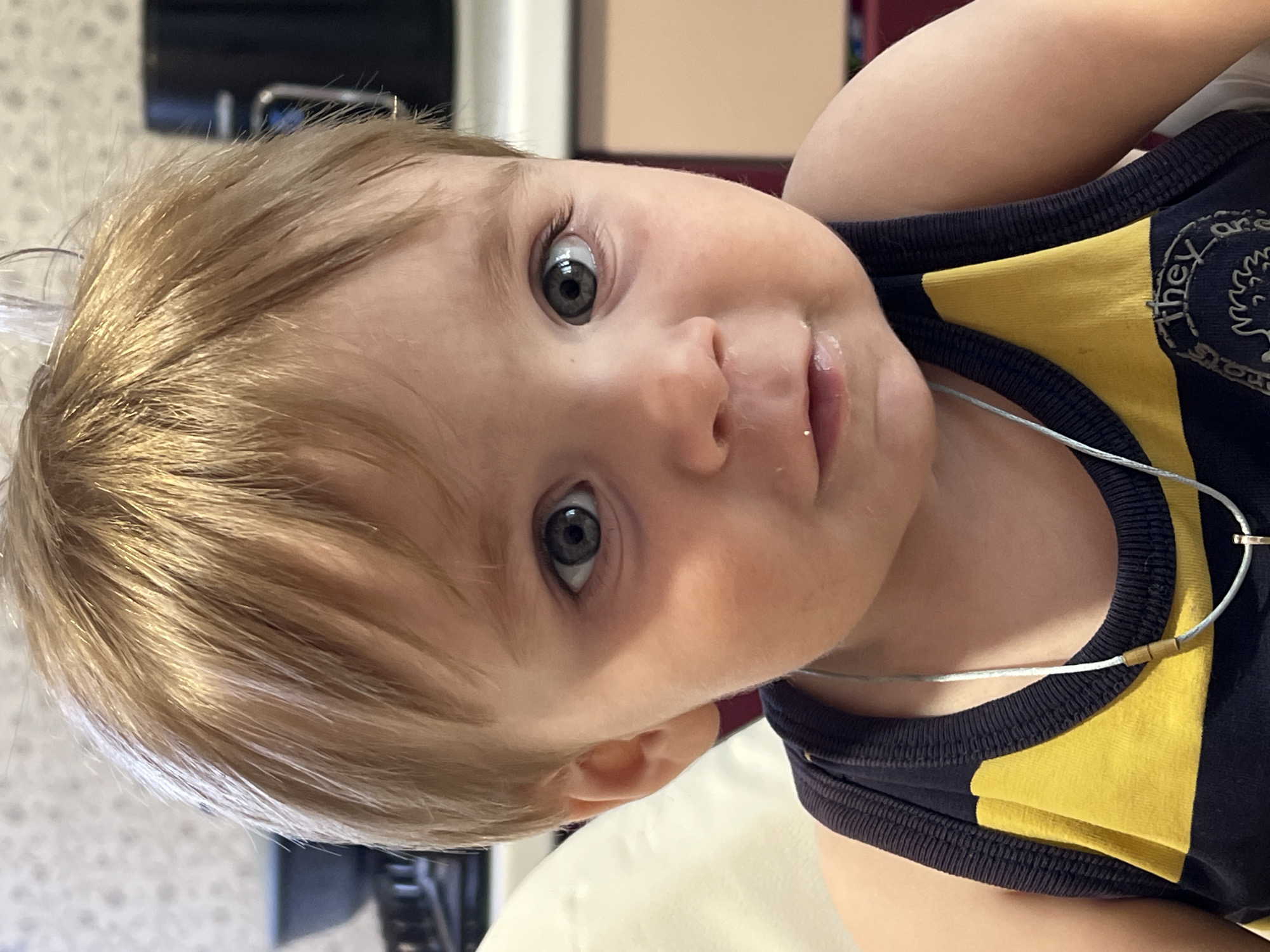 A close-up, medium shot depicts a fair-skinned infant with light blonde hair and gray-green eyes, wearing a navy and yellow tank top and a thin silver necklace. The child appears to be lying down, looking directly at the camera with a slight smile. A trace of liquid is visible on the upper lip, suggesting possible drool or food residue. The background is blurred and offers minimal detail, making it impossible to determine the exact location. There is no information about the location being in the city of Noviy, Russia in the picture provided.