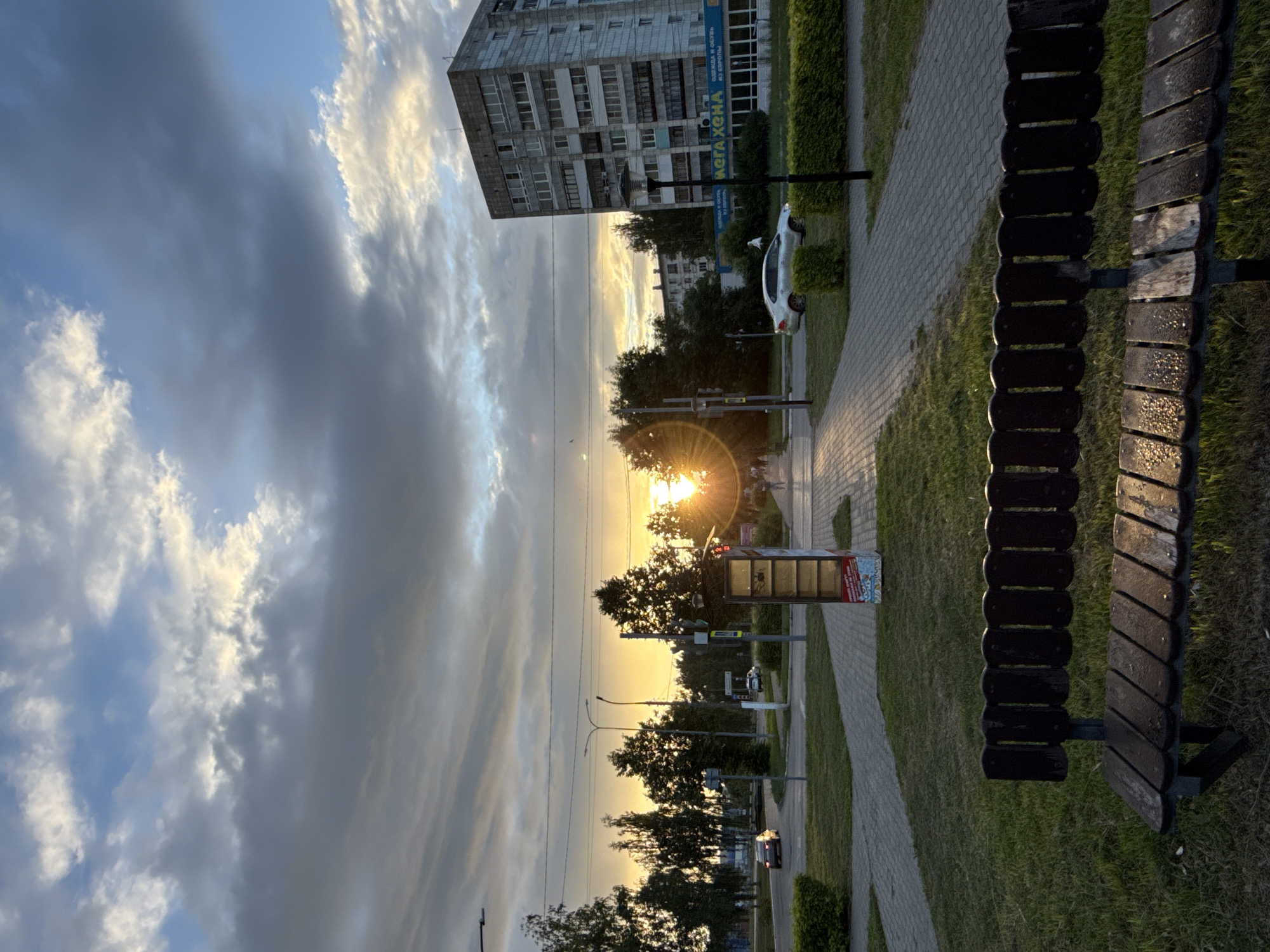 This media file depicts an urban street scene at sunset. The foreground features a weathered wooden bench, its slats showing signs of age and water droplets, suggesting recent rain or dew. The bench is positioned next to a paved walkway that curves alongside a grassy area. Beyond the walkway, trees line the street, with lush green foliage indicating a temperate climate.
In the mid-ground, a white car is parked on the side of the road, and a red and white kiosk or bus stop shelter stands near the walkway. Streetlights and power lines crisscross the scene. Further back, a multi-story apartment building with numerous windows dominates the skyline. The building's facade is weathered and utilitarian, typical of Soviet-era architecture.
The sky is filled with dramatic clouds, ranging from dark grey to bright white where illuminated by the setting sun. The sun itself is a prominent feature, casting a warm, golden glow and creating visible sunbeams. This indicates that the time of day is sunset, and the weather is likely clearing after rain, or is a generally partly cloudy evening.
The overall mood is one of peaceful, everyday life in a residential area, with the natural beauty of the sunset softening the urban landscape. The scene evokes a sense of quiet contemplation. No people are clearly visible in the image, adding to the tranquil atmosphere.
There is some visible text on the blue sign on the building, which appears to be Cyrillic, and text on the kiosk, also likely in Cyrillic. The text is too small and blurry to be reliably deciphered.