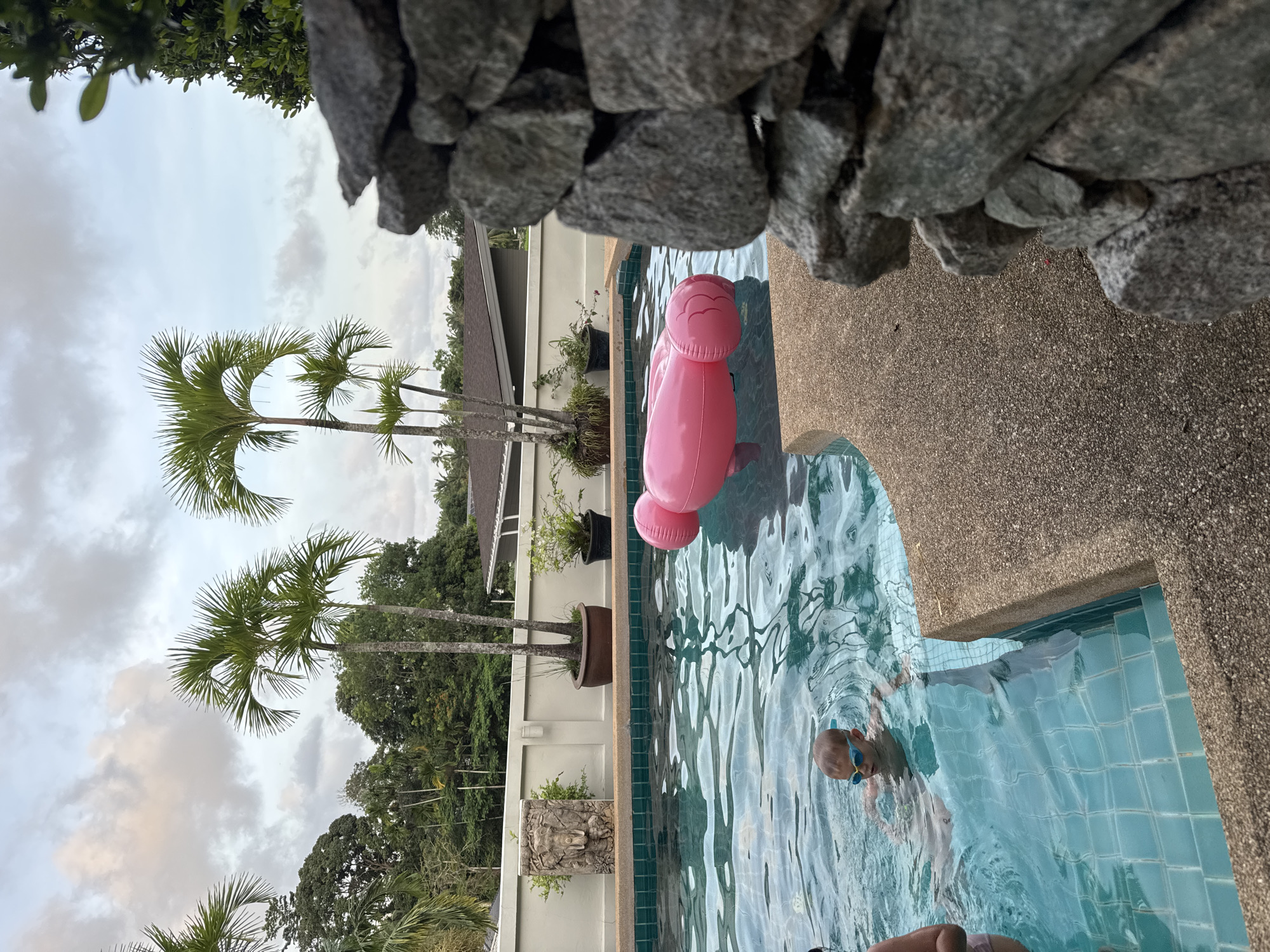 The image depicts a swimming pool located in Rawai, Thailand, next to a white wall and lush palm trees set against a cloudy sky. A child wearing blue goggles is in the pool. A pink inflatable toy floats in the water near the pool's edge. Rocks are visible at the top edge of the frame.