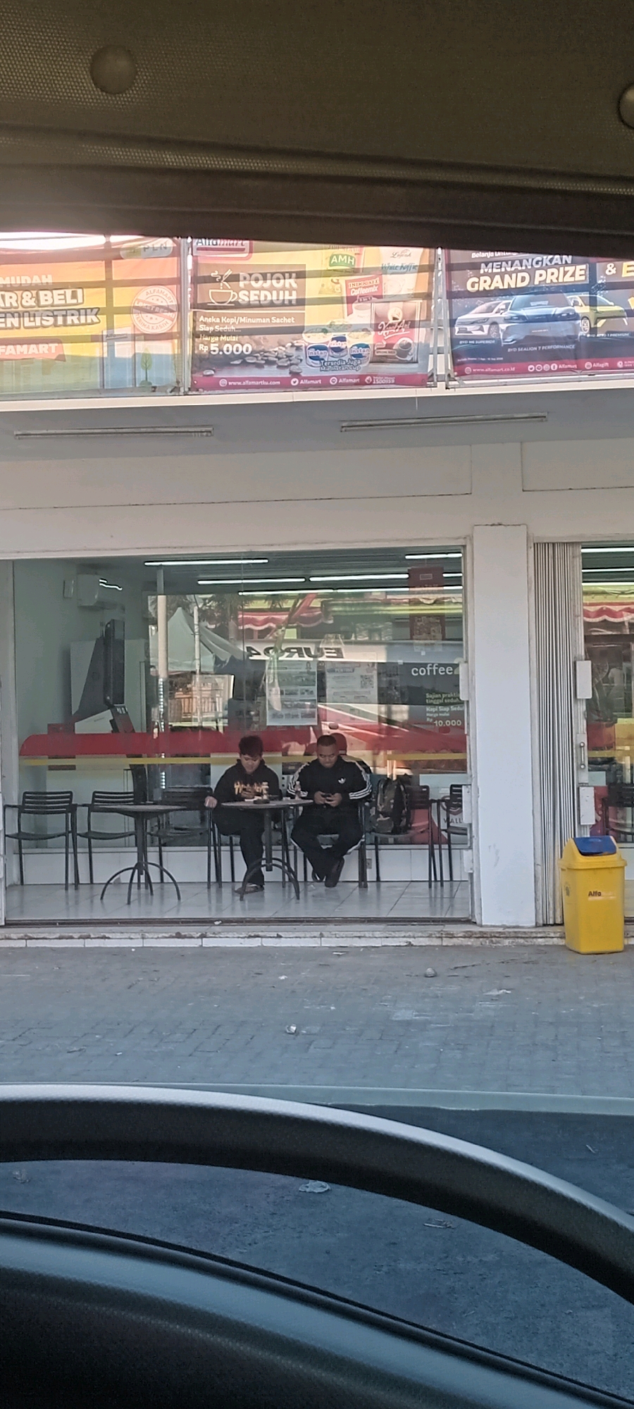 The image shows an outdoor seating area in front of a minimarket, possibly in Nguwok, Indonesia, judging by the signage. Two young men are seated at separate small tables, both looking at their phones. The minimarket has large glass windows, revealing shelves stocked with goods and what appears to be a refrigerator unit. Above the minimarket's entrance, there is a row of promotional banners. One banner displays an offer for "POJOK SEDUH" with a price of Rp 5.000. Another banner advertises "MENANGKAN GRAND PRIZE & E". To the right of the minimarket entrance, a yellow trash bin with a blue lid is visible. The ground in front of the minimarket is paved with interlocking bricks, and there are some scattered pieces of litter. The scene is captured from a slightly elevated perspective, possibly from inside a vehicle, as suggested by the presence of a car interior element in the foreground. The lighting indicates it is daytime, with shadows suggesting the sun is at an angle.