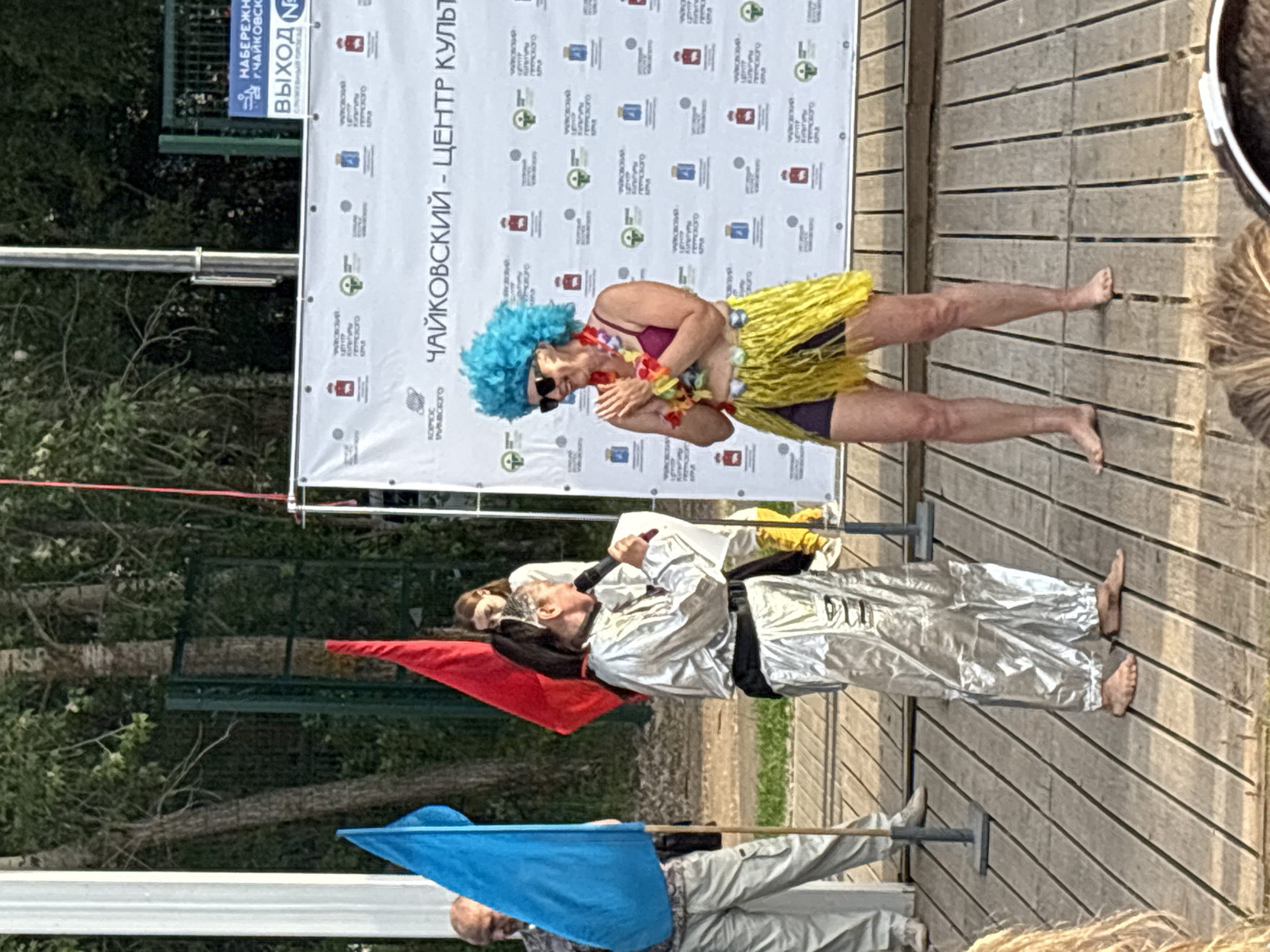 Two women are on a wooden stage in front of a banner. The woman on the left wears a shiny silver suit and is barefoot, holding a microphone and paper. The woman on the right wears a bright blue wig, sunglasses, a magenta top, and a yellow hula skirt. She has leis around her neck and her hands are clasped together. A blue flag and a red flag are behind the woman on the left. The banner behind them reads "Чайковский - Центр Культуры" which translates to "Chaykovsky - Center of Culture." The image appears to be taken at an outdoor event in Chaykovsky, Russia.