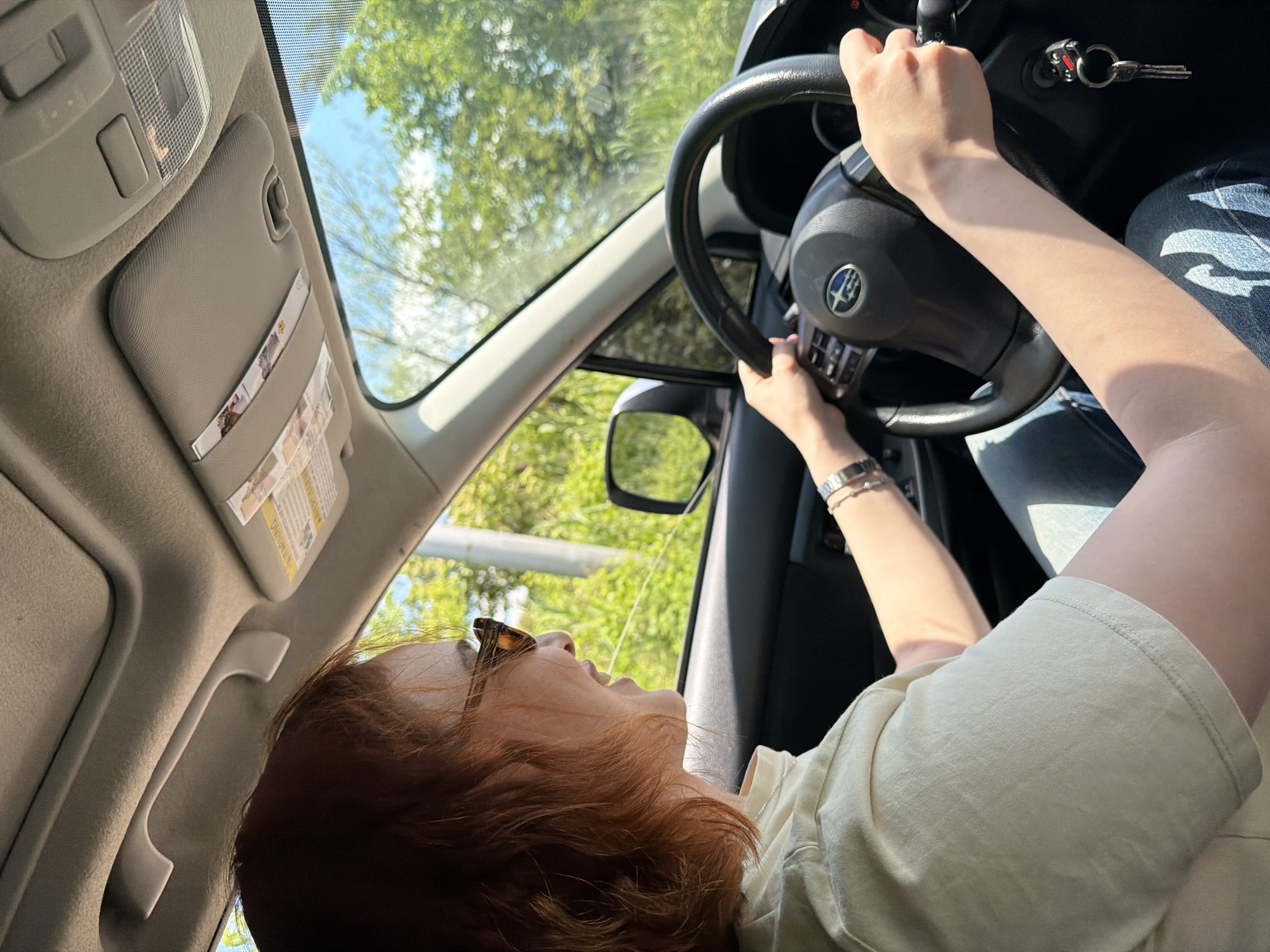 A woman with reddish hair is at the wheel of a car, presumably driving. She is wearing sunglasses and a light-colored shirt. Her hands are on the steering wheel, and a bracelet is visible on her left wrist. The car keys are in the ignition. The car's interior is visible, including the sun visor and rearview mirror. Outside the car, green trees and a blue sky are visible. There are no clear indicators that specify Tbilisi, Georgia.