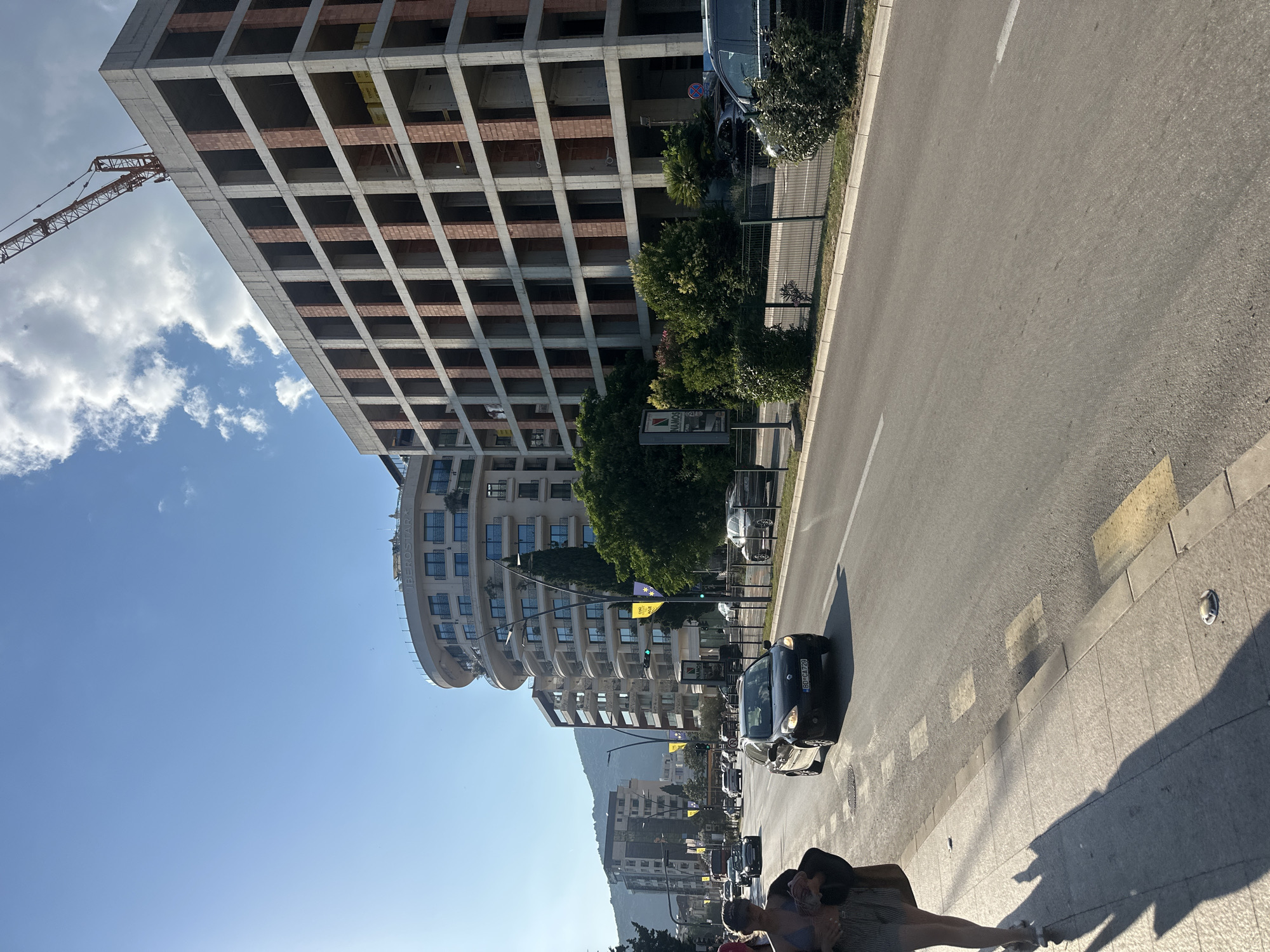 The image depicts a street scene in Budva, Montenegro, bathed in bright sunlight. In the foreground, a person walks on a sidewalk, their shadow stretching long across the pavement. The street is busy with vehicular traffic, including a dark-colored sedan in the middle lane and other cars further down the road. On the right side of the street, a large building under construction dominates the skyline, its concrete frame and brick infill visible. A construction crane extends from the top of this building. To the left of the construction site, a modern, circular-shaped building with balconies and a sign reading "IBEROSTAR" stands tall. Further in the background, other multi-story buildings line the street, and a lush green mountain slope is visible in the distance under a clear blue sky with scattered clouds. The environment appears to be urban, with well-maintained sidewalks, road markings, and streetlights.