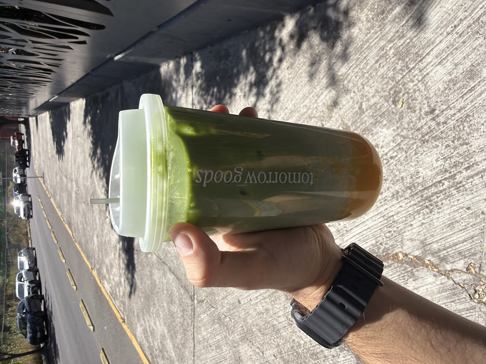 A close-up, high-angle outdoor shot, rotated 90 degrees clockwise, captures a person's right arm and hand holding a tall, clear plastic cup with a two-layered beverage. The drink features a vibrant green layer on top, likely matcha, and an orange/amber layer at the bottom, suggesting a mixed drink such as a matcha latte with a fruit base (e.g., mango or orange). The brand "tomorrow goods" is visible in white, inverted text on the side of the cup. A translucent white lid with a slot for a straw, and a small white straw inserted, complete the drink's presentation.
The hand, with light to medium skin tone and some visible arm hair, grips the cup firmly. A black, ribbed smart watch or fitness tracker is worn on the wrist.
The background shows a sunlit outdoor environment. Below the arm, a concrete or asphalt surface with a rough texture is visible, suggesting a sidewalk or paved area. Further back, a road stretches into the distance with a yellow painted line marking a lane or parking area. Several cars, mostly light-colored, are parked along the side of the road. On the left side of the frame, there's a dark, ornate metal fence or decorative wall with intricate cut-out patterns, casting strong, elongated shadows across the ground, indicative of bright midday or early afternoon sun. The strong sunlight creates clear, distinct shadows from both the fence and the arm/cup. The overall scene suggests an urban or suburban setting in Puebla, Mexico, on a clear, sunny day.