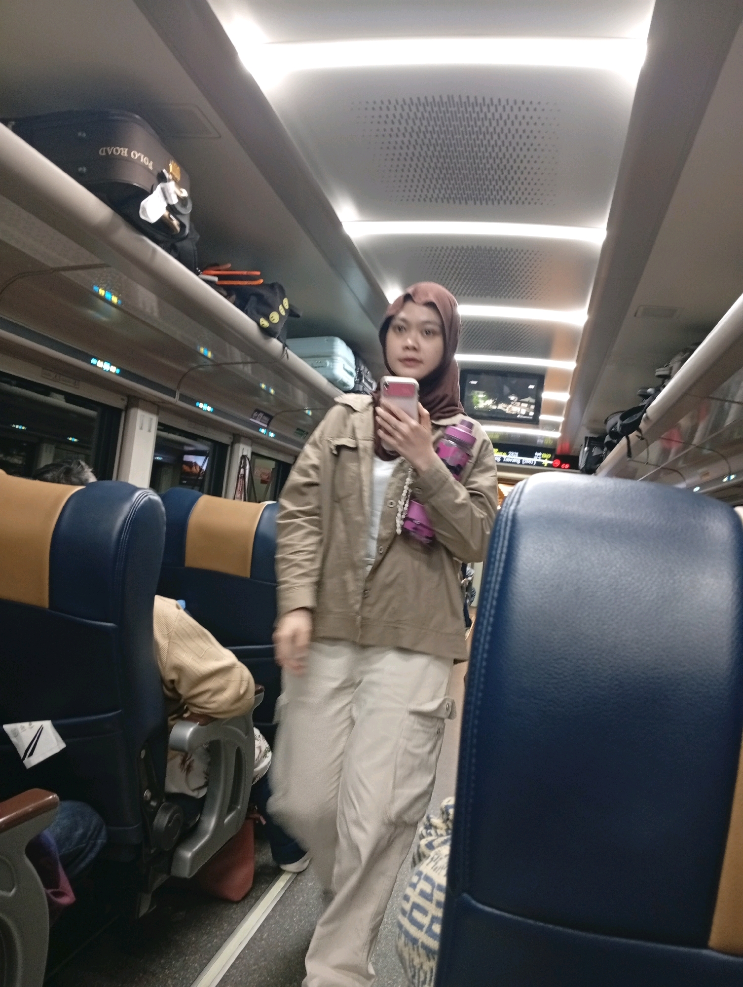 This media file captures a medium shot of a young woman standing inside a train carriage, holding a pink smartphone. She is wearing a brown hijab and a light brown jacket over a white top, paired with loose-fitting beige pants. Around her neck, she has a beaded necklace and two pink water bottles are attached to her jacket. The train carriage appears to be modern, with overhead compartments filled with luggage. One suitcase in the compartment above her clearly displays "POLO ROAD" in white lettering. The seats are upholstered in blue and tan, and the lighting suggests it might be evening or a dimly lit interior. Other passengers are visible in the background, with their backs to the camera, seated. The overall mood is casual and suggests a journey in progress. The location is Astanalanggar, Indonesia.