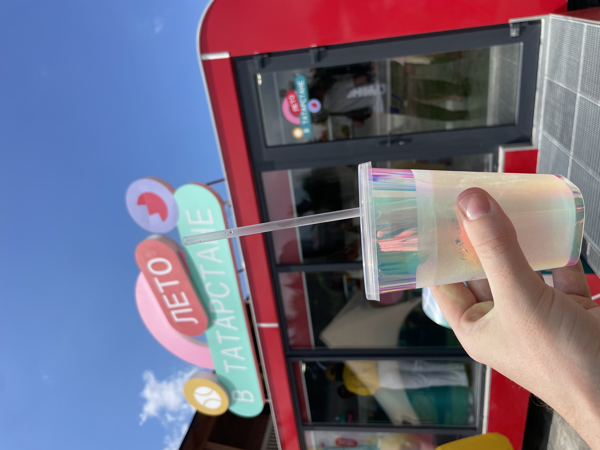 In Kazan, Russia, a hand holds a shimmering iridescent plastic cup filled with a pale, translucent beverage and a clear plastic straw. The cup is positioned in the foreground, obscuring a portion of the scene. In the background, a bright red, kiosk-like structure with large glass windows is visible. Above the kiosk, a sign reads "ЛЕТО" (Summer) in red lettering within a pink shape, and "В ТАТАРСТАНЕ" (In Tatarstan) in white lettering on a teal oval. The sky above is a clear blue with scattered white clouds. Inside the kiosk, blurry figures can be seen, one wearing a yellow top and white pants, and another in a light-colored shirt. The overall scene suggests an outdoor location, likely a summer event or market, in Kazan.
