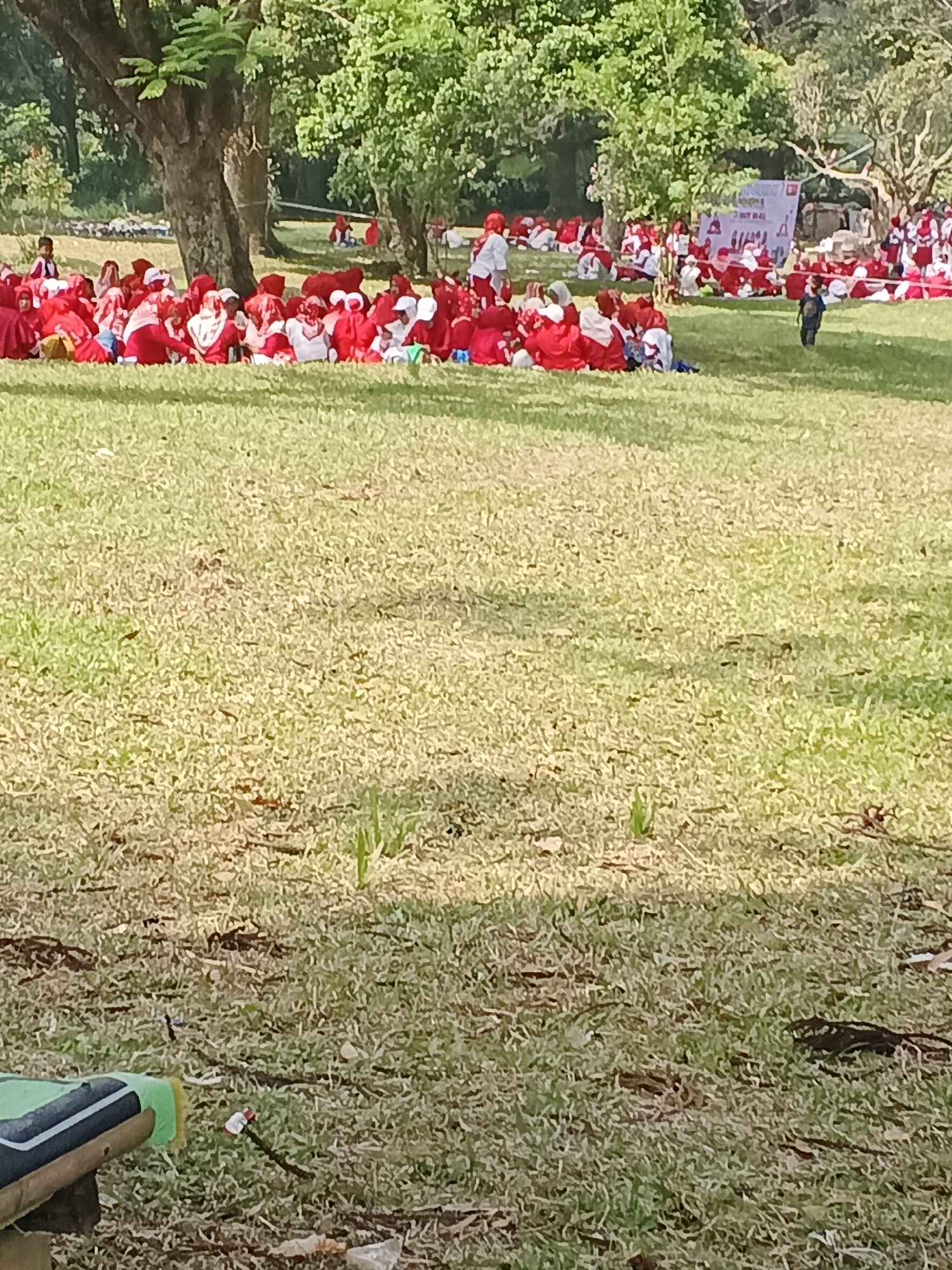 A crowd of people, many wearing red and white clothing and head coverings, are sitting on a grassy field. Some of them appear to be children. In the background, there are trees and a banner with writing on it. A small child in dark clothing is walking away from the camera towards the right side of the frame. The ground is covered in dry grass with some scattered leaves and debris. The image appears to be taken in an outdoor setting during the daytime.