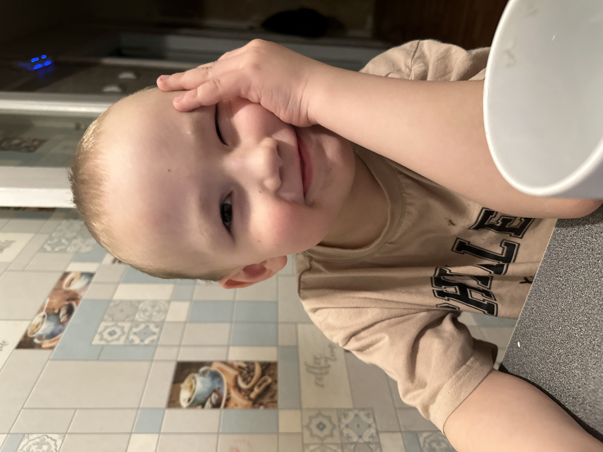 An indoor, medium shot captured in Chaikovsky, Russia, shows a young child looking towards the camera from a low angle, appearing to rest on a dark countertop. The child has short, light blonde hair and fair skin, with a slightly rosy complexion on the cheeks. One hand is placed playfully on their forehead and side of the face, covering part of their eye, while the other arm rests on the dark, flecked countertop. The child is wearing a light brown or beige t-shirt with dark, possibly collegiate-style letters visible on the chest, though the full text is unclear. There might be some residue or faint smudges around the child's mouth.
To the right, a white cup is partially visible, suggesting it's on the countertop near the child. The floor or lower wall in the background features light-colored rectangular tiles. Some tiles have intricate light blue and white geometric patterns, while others depict illustrations of coffee cups with beans and a script font spelling out "coffee." In the darker background, out of focus, a series of small blue indicator lights are visible, possibly from an electronic device or appliance. The overall lighting suggests an indoor setting, possibly in the evening or under ambient artificial light. The child's expression is one of slight coyness or a gentle smile, indicating a comfortable and playful mood.