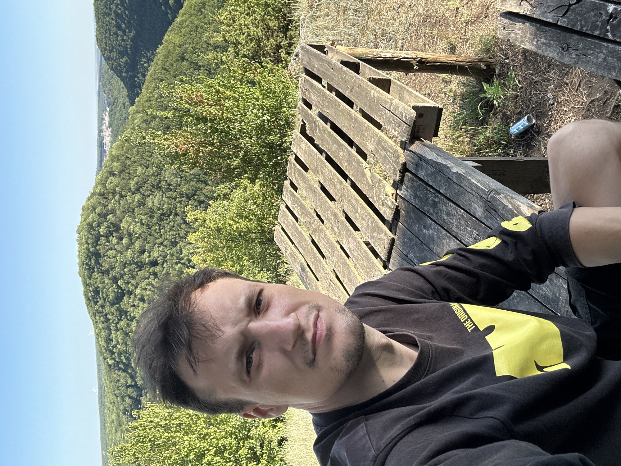 A man is taking a selfie in an outdoor setting with a hilly, forested landscape in the background. He is wearing a black long-sleeved shirt with a large yellow graphic on the front and yellow text on the sleeve. He is seated next to a weathered wooden picnic table, which appears to be constructed from pallets. The ground around the table is covered in dry grass and dirt. A blue and silver can is visible on the ground near the table leg. The sky is clear and blue, and the sun is shining, casting shadows. The location is identified as Pryvorottia Druhe, Ukraine.