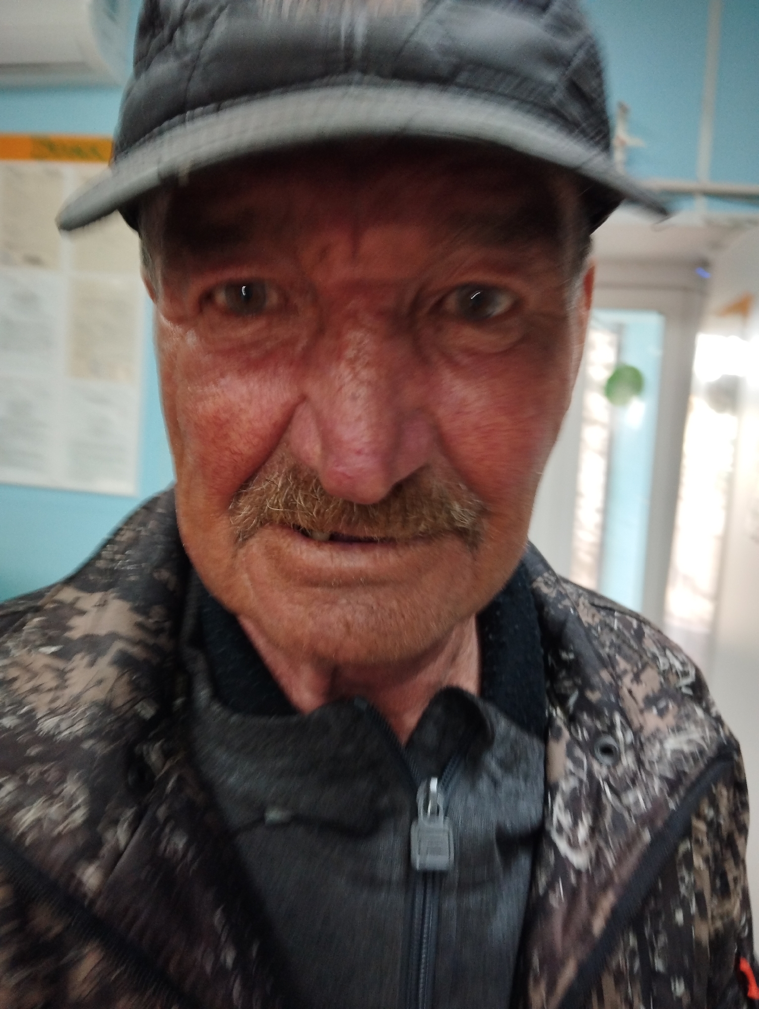 A close-up shot of an elderly Caucasian male wearing a dark-colored cap and a camouflage jacket with a dark undershirt or sweater visible. The man has a mustache. The background shows a wall with a notice board. The location is in a room, likely indoors, in Krasnogorskoye, Russia. A door frame and window are faintly visible in the background.