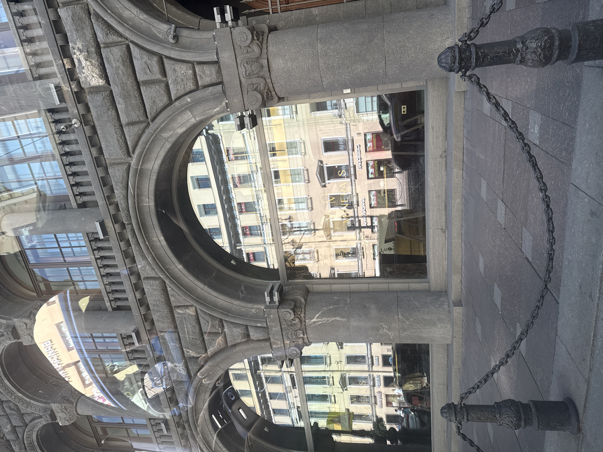 This is a medium-shot, eye-level, outdoor photograph of the reflection in a large glass window of a stone building in Saint Petersburg, Russia. The building's facade features a series of arched openings with decorative stonework. The reflection shows a street scene with several buildings across the street. These buildings are multi-story and have numerous windows. Some of the buildings appear to be commercial, with signs visible in Russian. A dark-colored car is parked on the street, and several people can be seen walking. In the foreground, a dark metal bollard with a decorative design is visible, connected by a dark metal chain to another bollard to the right. The ground is paved with square and rectangular stones. The overall impression is of an urban environment with a mix of historical and modern elements.