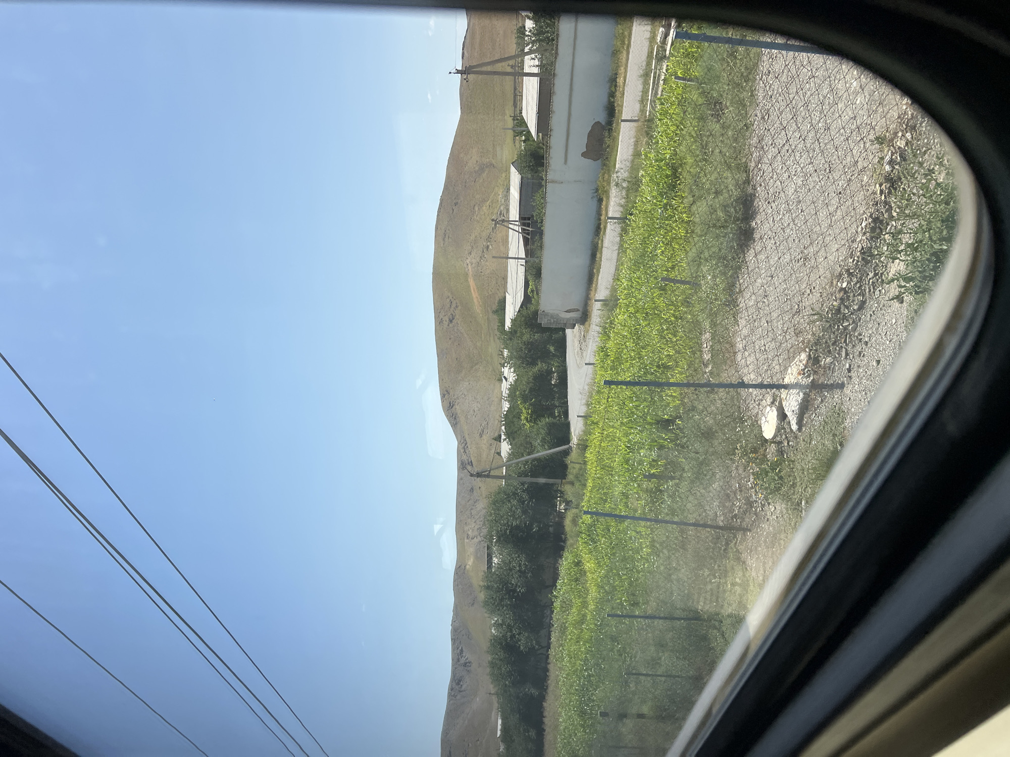 The image depicts a rural scene viewed through a curved window, likely from a vehicle. The dominant subjects are a dry, barren hillside on the left and a lush, green field on the right. The hillside is sculpted and devoid of significant vegetation, suggesting an arid climate. In contrast, the field is vibrantly green and appears to be a cultivated crop, possibly corn or similar, with rows clearly visible.
Between the hillside and the field, several man-made structures are present. There are white or light-colored buildings with corrugated roofs, suggesting agricultural outbuildings or simple dwellings. Power lines are visible running across the sky and also along the edge of the field, supported by utility poles. A chain-link fence runs along the right side of the image, separating the field from a gravel path or road.
The sky is a clear, bright blue, indicating a sunny day. The time of day appears to be midday or early afternoon, given the bright sunlight and lack of long shadows. The overall atmosphere is one of rural simplicity and agricultural activity, set against a backdrop of natural, rugged terrain.
There is no visible text in the image. The scene is characteristic of a location in Almachi, Uzbekistan, with its dry hills and agricultural fields. The image captures a moment of travel, with the curved window framing the passing landscape.