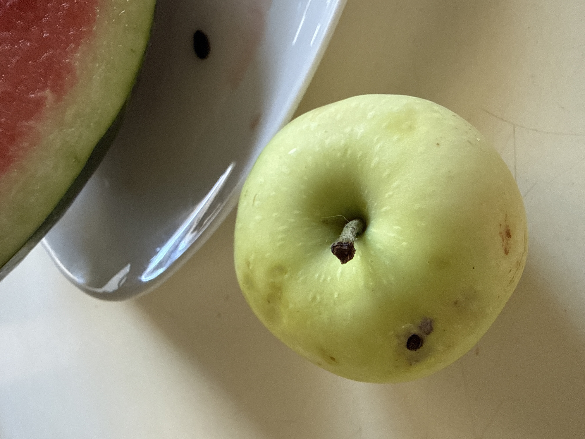 A close-up, overhead shot features a light green apple with a brown stem. The apple is positioned on a light-colored surface, possibly a table. In the background, a white plate holds a slice of watermelon. The scene does not depict any specific location in Okhtyrka, Ukraine, or any people, actions, or interactions. The environment is neutral and appears to be indoors.