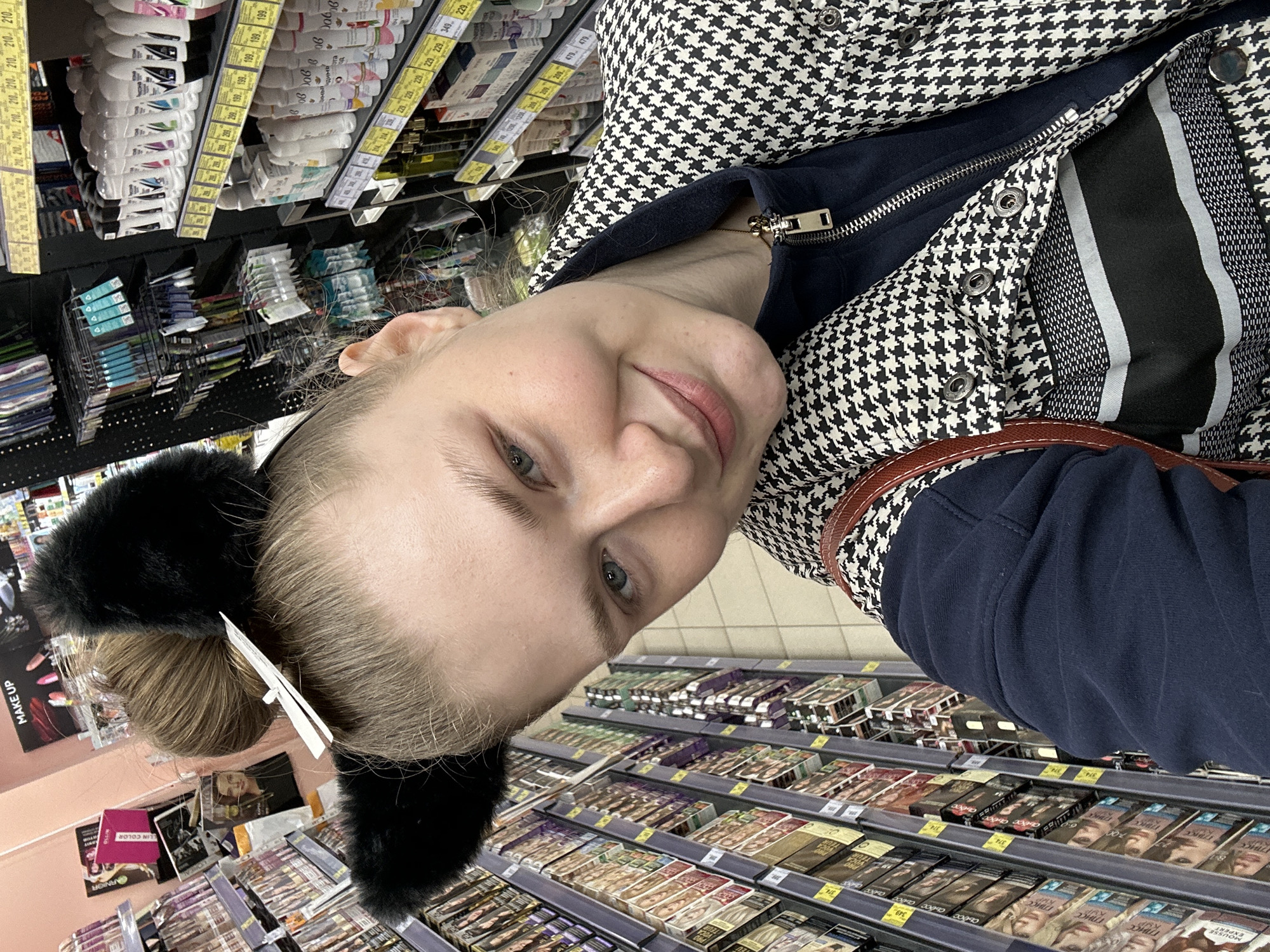 A woman is taking a selfie inside a store, likely a cosmetic or general store. She has light skin, blue eyes, and her hair is pulled back with a black headband featuring furry ears. She is wearing a houndstooth pattern jacket over a dark blue top. A brown strap from a bag or purse is visible over her shoulder. The shelves behind her are stocked with various hair dyes and cosmetics. Above her are shelves with personal hygiene products. Price tags are visible on the shelves. The store is located in Kazan, Russia, as indicated by the product packaging written in Russian.