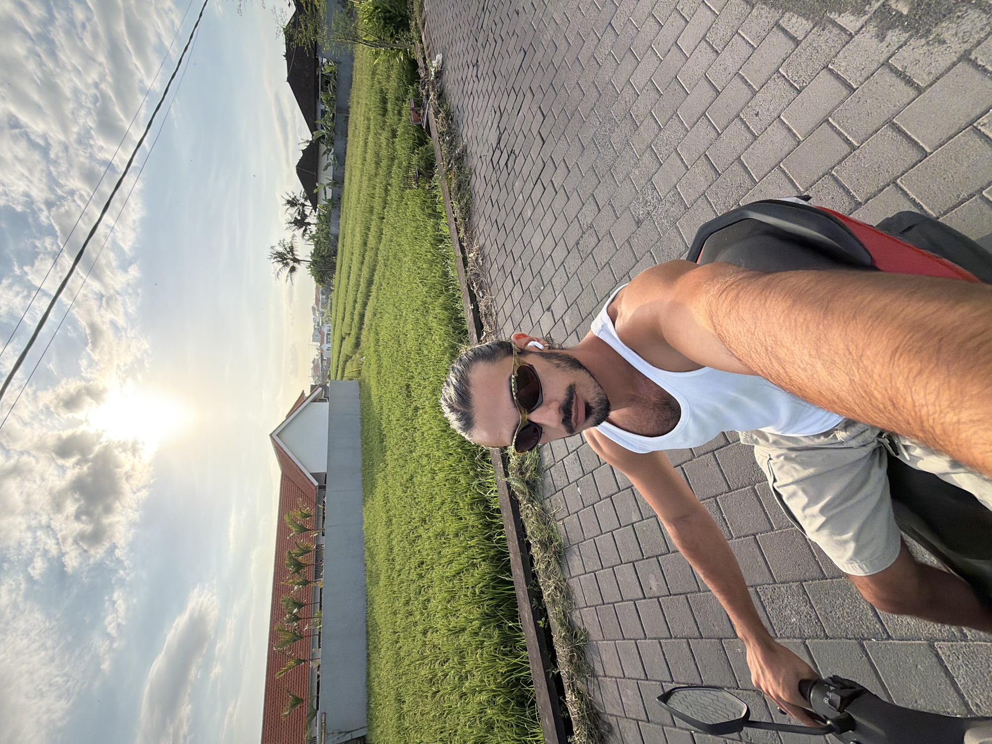 A man wearing a white tank top, tan shorts, and sunglasses is riding a motorcycle on a brick-paved road in what appears to be a rural area. The road is flanked by a lush green rice field on one side and buildings on the other. The sky is visible in the background, suggesting it is daytime. Based on the location in the city of North Kuta, Indonesia, it can be assumed that the rice field is typical of the landscape in that area.