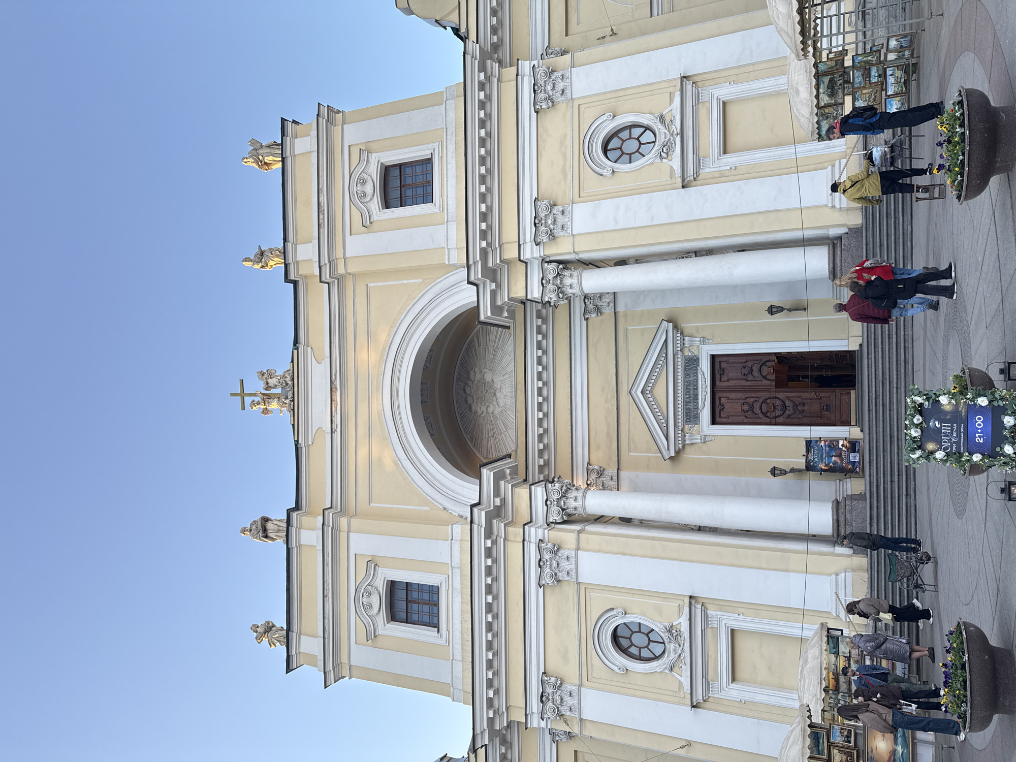 The image shows the facade of Saint Catherine's Church in Saint Petersburg, Russia, angled upwards from ground level. The church has a cream-colored facade with white architectural details, including columns, window frames, and decorative elements. There are statues atop the church. The facade includes an arched opening above the main entrance, and a cross on top of the building. The main doors are visible. Several people are standing and walking on the steps and pavement in front of the church. Some appear to be looking at artworks displayed on stands. A decorated pot containing flowers is visible in the foreground. The sky is clear and blue. A sign displays the time 21:00.