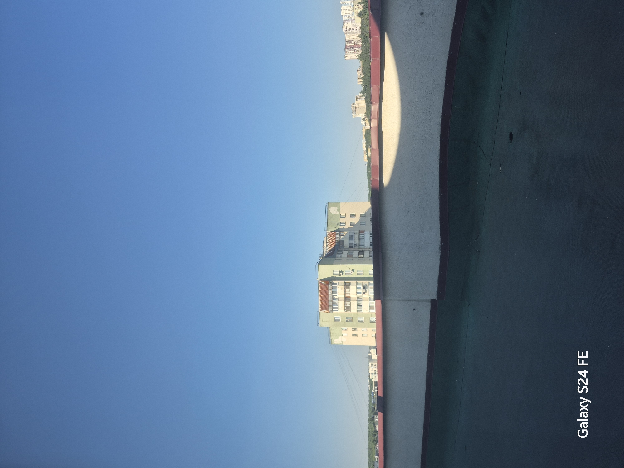 A wide shot captures a clear blue sky above a cityscape. In the mid-ground, a multi-story apartment building, painted in light green and peach tones with a reddish-brown roof, stands prominently. It features numerous windows and balconies. To the right, other apartment buildings are visible in the distance, and a line of trees separates them from the closer structures. In the foreground, a rooftop surface with a textured, dark green material is visible, bordered by a light-colored wall with a dark trim. A large, semi-circular shadow falls across this wall, cast by an unseen object. The bottom left corner of the image displays the text "Galaxy S24 FE". The scene appears to be a rooftop view in Chișinău, Moldova, likely during the daytime due to the bright sky and shadows.