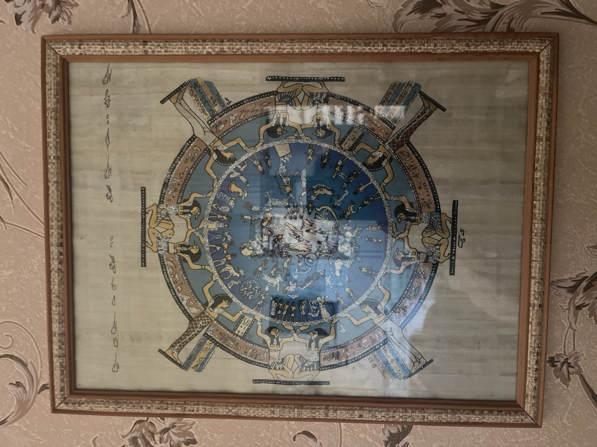 This is a framed Egyptian papyrus art piece depicting a zodiac or astronomical chart, hanging on a patterned wallpaper wall. The artwork is circular with figures and hieroglyphs arranged around a central blue area. The frame is made of wood with a woven texture. The background wallpaper has a floral or abstract pattern in shades of beige and pink. The scene is an indoor shot, presumably in a home. The artwork's content suggests a historical or astrological theme. There are no visible people or specific actions taking place within the image itself, beyond what is depicted in the artwork. The location of Vladimirovka, Russia is not identifiable from the visual information in the image.