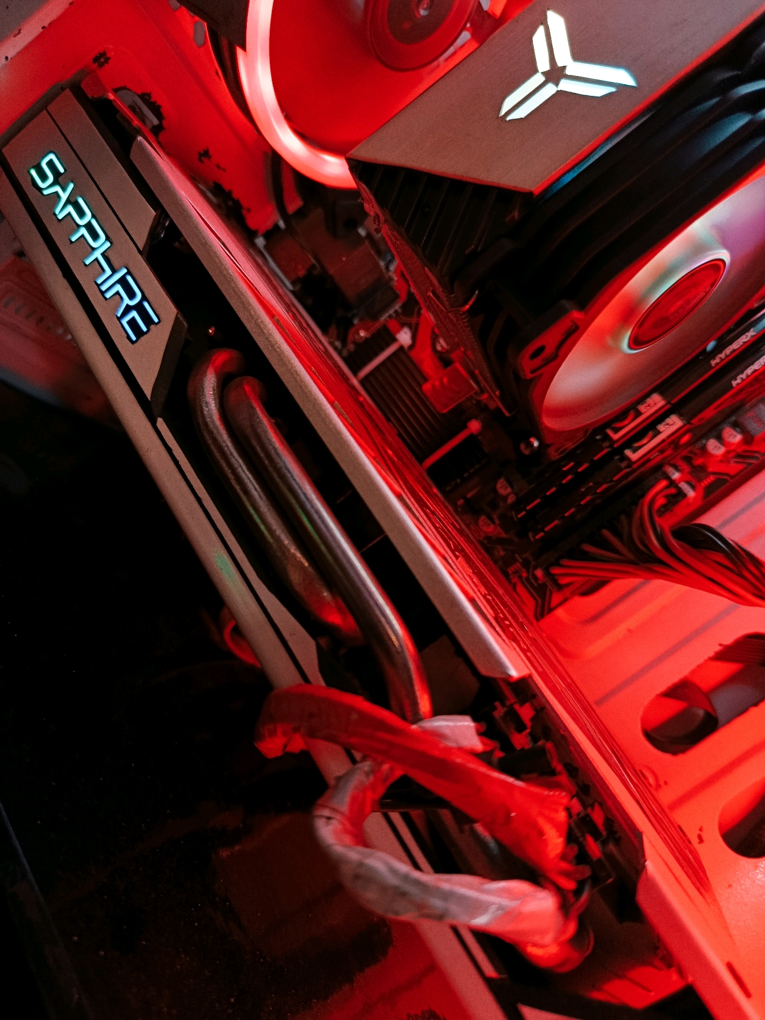 This is a close-up, high-angle, tilted indoor shot of the interior of a computer tower, illuminated by red lighting. The primary subject is a graphics card with "SAPPHIRE" written on it in glowing blue letters. The graphics card has prominent metal heat pipes. To the right of the graphics card, a portion of the motherboard is visible, with RAM modules installed. Above the graphics card, a component with a stylized white logo, resembling an asterisk or a fan, is lit up. The overall scene is characterized by red ambient lighting, which casts a glow on the internal components of the computer. There are no people present, and the environment is the inside of a computer case.