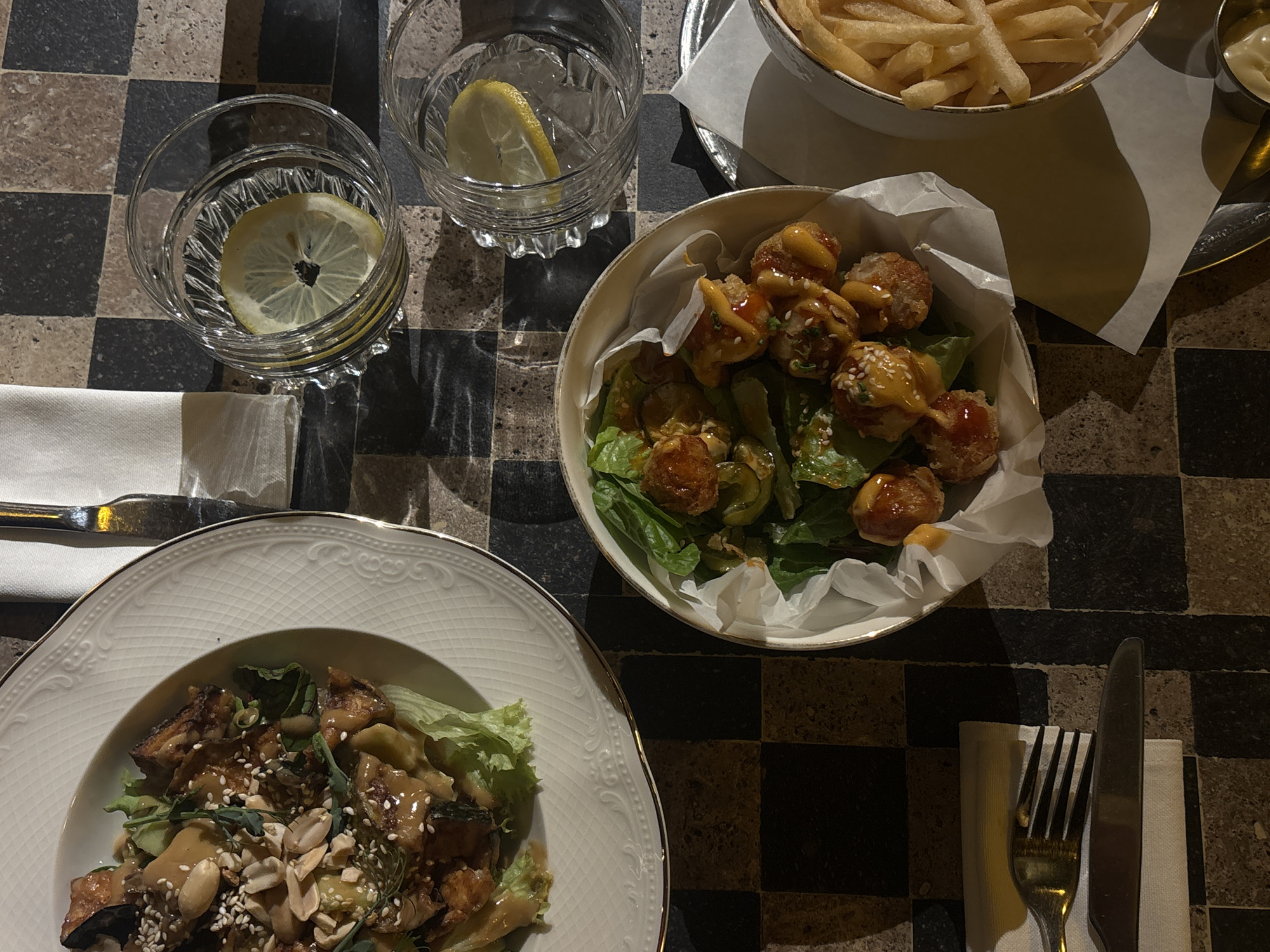 The image provides an overhead view of a dining table in what appears to be a restaurant setting in Yerevan, Armenia. The table features a distinctive dark checkered pattern, with alternating black and dark grey/brown squares.
Several dishes and drinks are arranged on the table. Two clear glasses of water are visible, each containing a lemon slice; one glass also contains ice cubes. Food items include a bowl filled with golden-brown french fries, served alongside a small metal dish holding a white dipping sauce, likely mayonnaise. Another bowl contains what appears to be fried appetizer portions, possibly chicken or shrimp, coated in a creamy orange sauce and sprinkled with sesame seeds, presented on a bed of green lettuce with some green pepper pieces. A third dish is a white plate with an ornate, gold-rimmed edge, holding a salad composed of green lettuce, chunks of what seems to be roasted vegetables or protein, peanuts, and sesame seeds, all drizzled with a light brown, creamy dressing.
Cutlery, including forks and knives, is placed on white napkins beside the plates. The lighting is dim, creating noticeable shadows, which suggests an evening or nighttime atmosphere. No people are visible in the frame, and there is no discernible text.