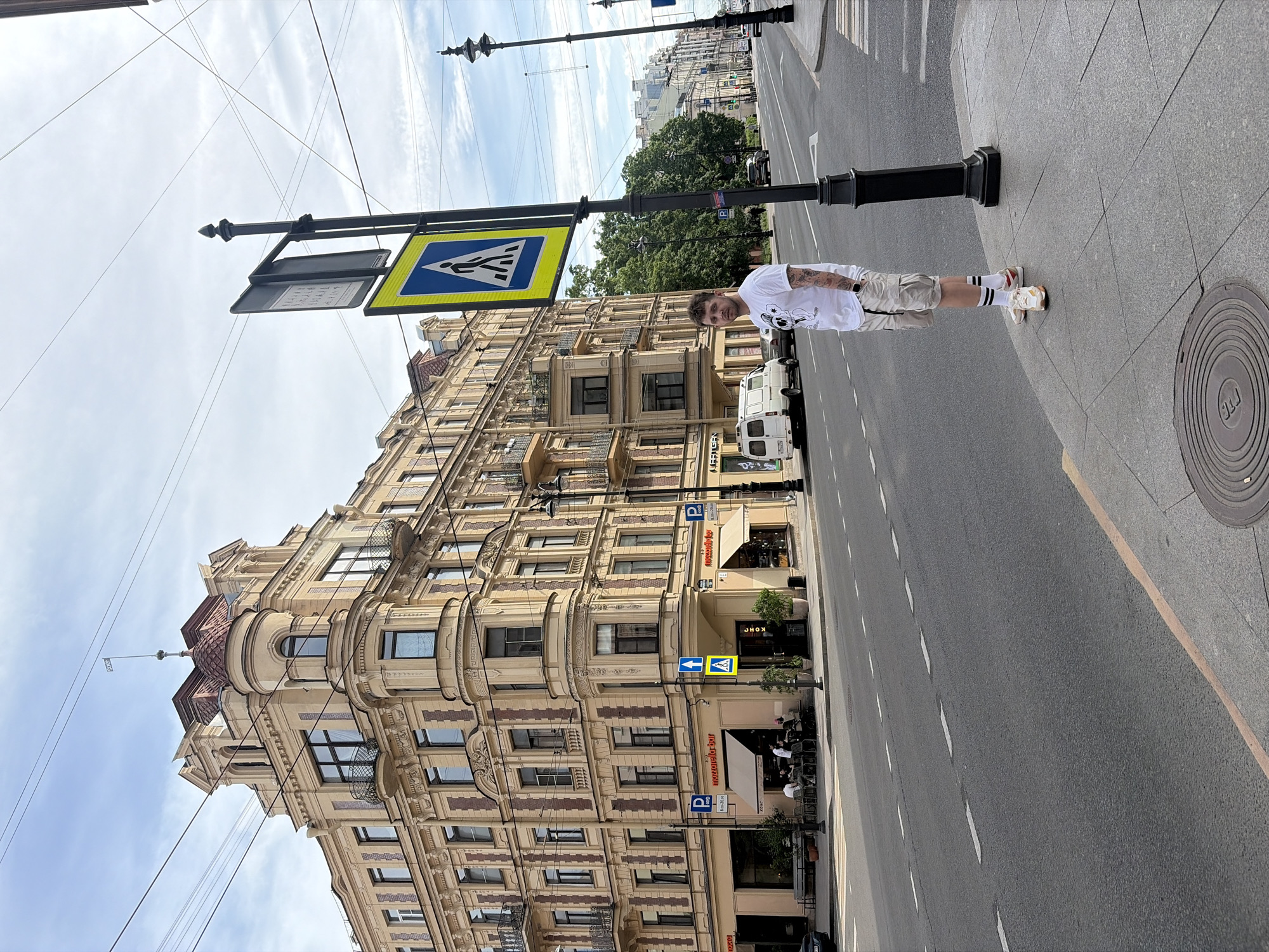 The image depicts a street scene in Saint Petersburg, Russia, specifically at the corner of Malaya Konyushennaya Ulitsa and Bolshaya Konyushennaya Ulitsa, where the building at Malaya Konyushennaya Ulitsa, 1/3A is located.
The prominent structure on the left is an ornate, multi-story corner building, light yellow in color with reddish-brown decorative brickwork on its facades. It features multiple windows, several balconies with wrought-iron railings, and a rounded bay window that culminates in a red-domed turret topped with a spire. A small white rectangular flag-like sign bearing the text "SOHO" is affixed to the spire. The ground floor of the building houses businesses, including "Mozzarella Bar" and "KONO" (КОНО), both with awnings and outdoor seating areas where people are visible. Two blue "P" parking signs indicating hours "8-20:00" are also present.
In the right foreground, a fair-skinned male individual stands on a gray tiled sidewalk, positioned next to a black lamppost. He has short brown hair, a beard, and visible tattoos on his forearms. He is wearing a white t-shirt with a graphic print, light beige cargo shorts, white socks with black stripes, and white and orange sneakers. He looks directly forward.
The lamppost next to the man displays a blue square sign with a white walking figure, indicating a pedestrian crossing. Above it is a smaller, dark rectangular sign. The asphalt road in front of the building has white dashed lane markings and a yellow curb line on the right side. A pedestrian crosswalk is visible on the street. A circular metal manhole cover with Cyrillic text is present on the sidewalk in the lower right. In the mid-ground, a white van and other vehicles are parked or moving along the street, which is lined with green trees further down. Overhead power lines crisscross the blue sky, which has scattered white clouds. Another black lamppost extends further down the street on the right.