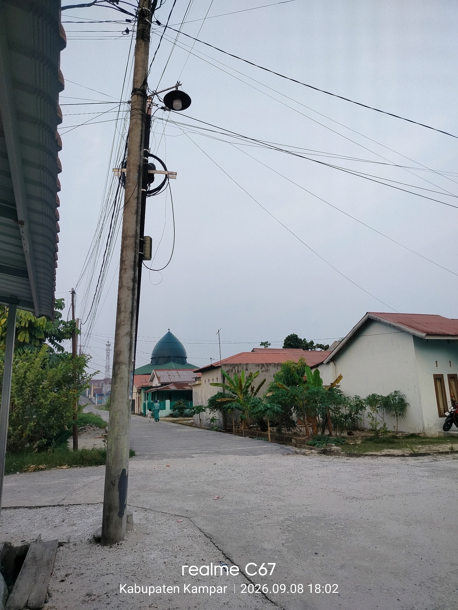 The image captures a street scene in Kubang Jaya, Indonesia, under an overcast sky. A prominent concrete utility pole, laden with a dense network of electrical wires and a hanging lamp, stands on the left side of the frame. To the left of the pole, a portion of a corrugated metal roof structure is visible. Further down the street, a green-domed mosque is the focal point, with several residential buildings flanking it. One house on the right is painted white with a reddish-brown roof and dark-framed windows. Two individuals, appearing to be women in headscarves, are walking away from the viewer towards the mosque. Lush green vegetation, including banana trees, is interspersed among the buildings. The road surface is paved with asphalt and gravel. The bottom right corner of the image displays a watermark indicating "realme C67" and the location and date "Kabupaten Kampar | 2026.09.08 18:02".