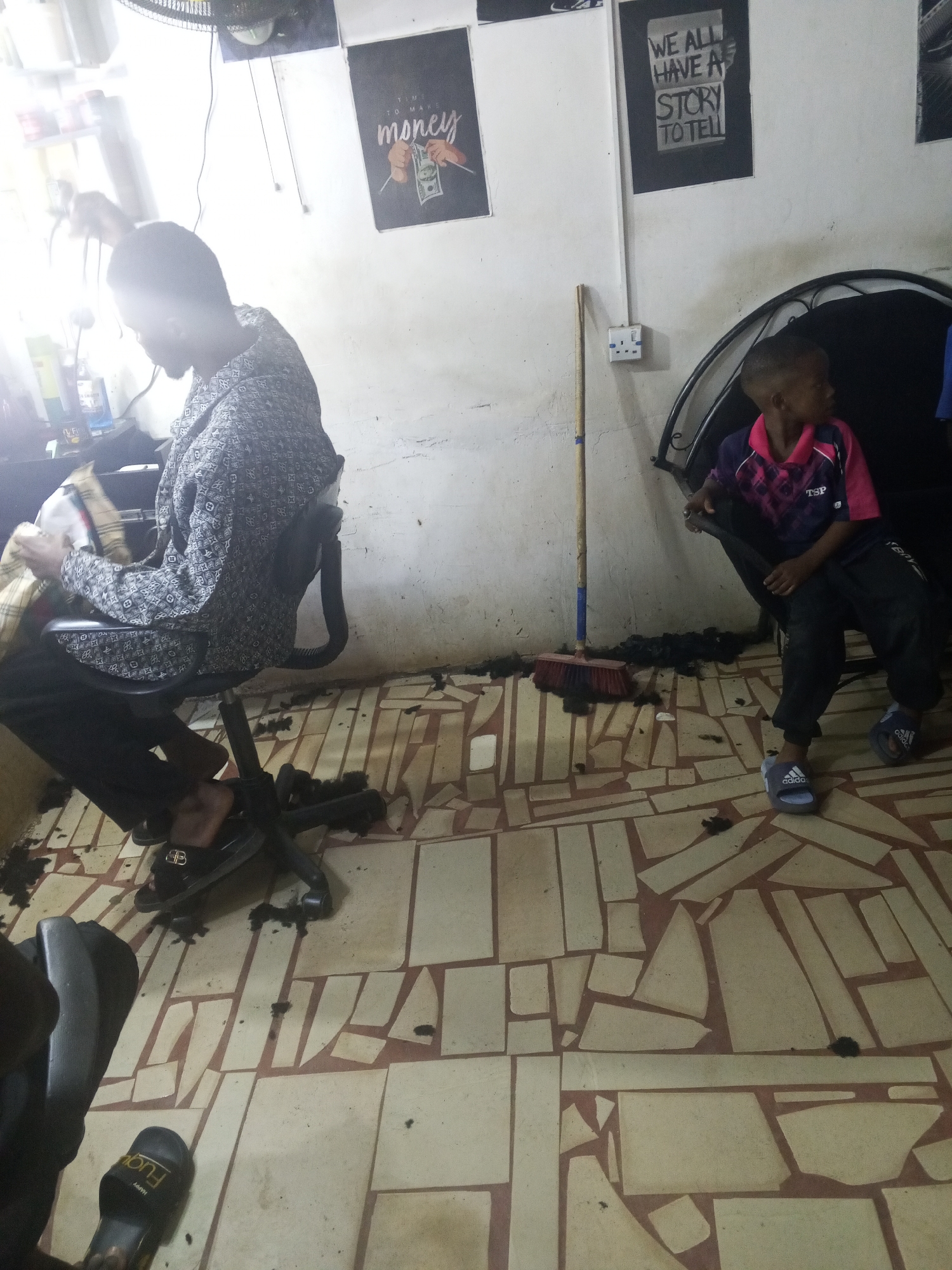 The image captures an interior scene, likely a barbershop, in what is possibly Gwong, Nigeria. A person is seated in a black office chair getting a haircut. Hair clippings are scattered on the floor. To the right, a child wearing a pink and purple shirt with black pants and blue Adidas slides sits in a dark chair with an arched back. The walls are painted in an off-white color. A broom leans against the wall near an electrical outlet. There are two framed posters on the wall. One poster reads "Time to make money" and depicts hands holding money. The other poster reads "We all have a story to tell." The floor is tiled with irregular shapes in light tan and brown.