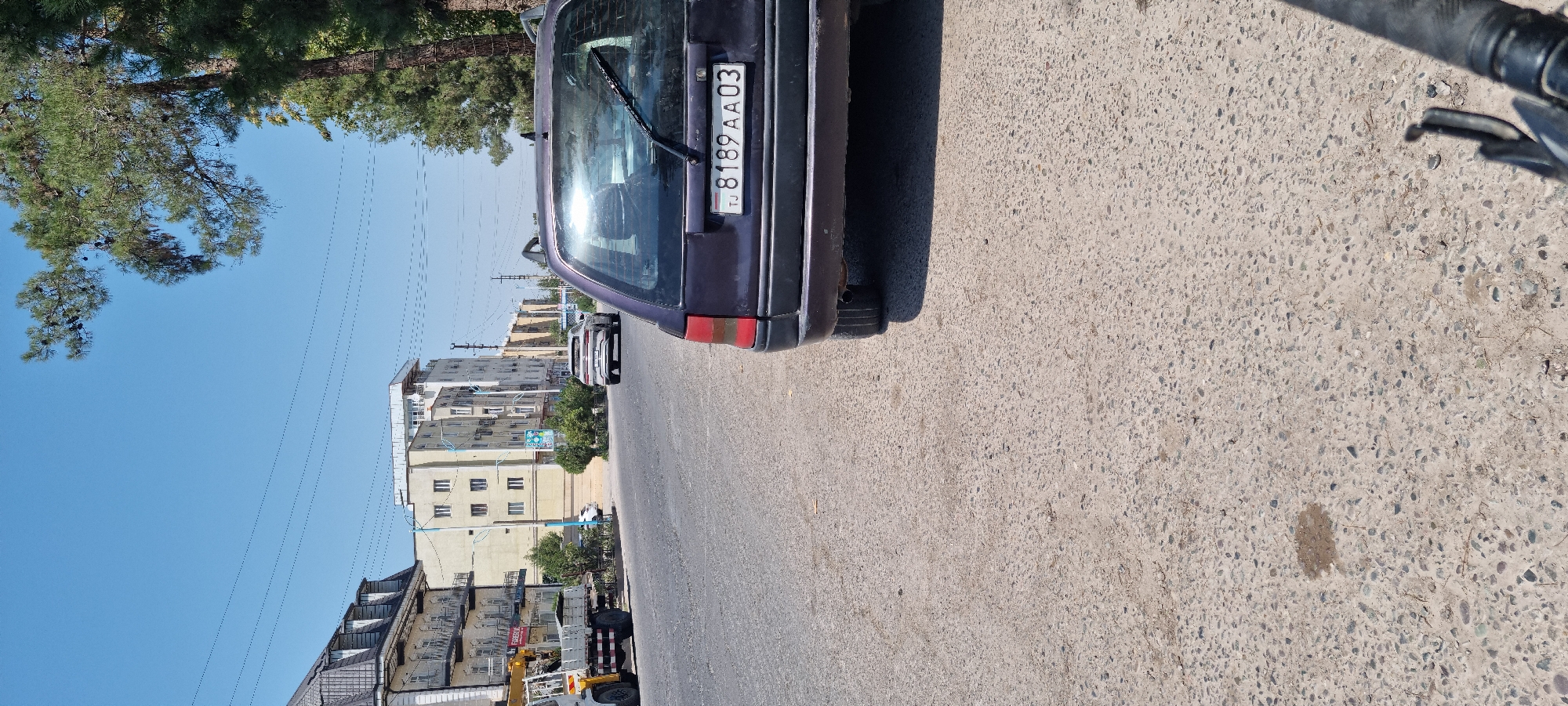 In the city of Ismoili Somoni, Tajikistan, on a sunny day, a dark purple hatchback is parked on the right side of a street. The car's rear is visible, including its license plate which reads "TJ 8189 AA 03". The street is paved with asphalt and has a slightly textured surface. To the left of the car, a street stretches towards the horizon, lined with multi-story buildings. One building, on the far left, features a dark roof and multiple windows. Further down the street, another building appears lighter in color with several floors and windows. Across the street, more buildings and trees are visible. Power lines are strung across the sky, supported by poles. A construction vehicle with a crane is parked on the left side of the street, near the buildings. In the distance, another white vehicle is seen driving on the street. The environment appears to be urban, with a clear blue sky overhead and mature trees lining the right side of the road.