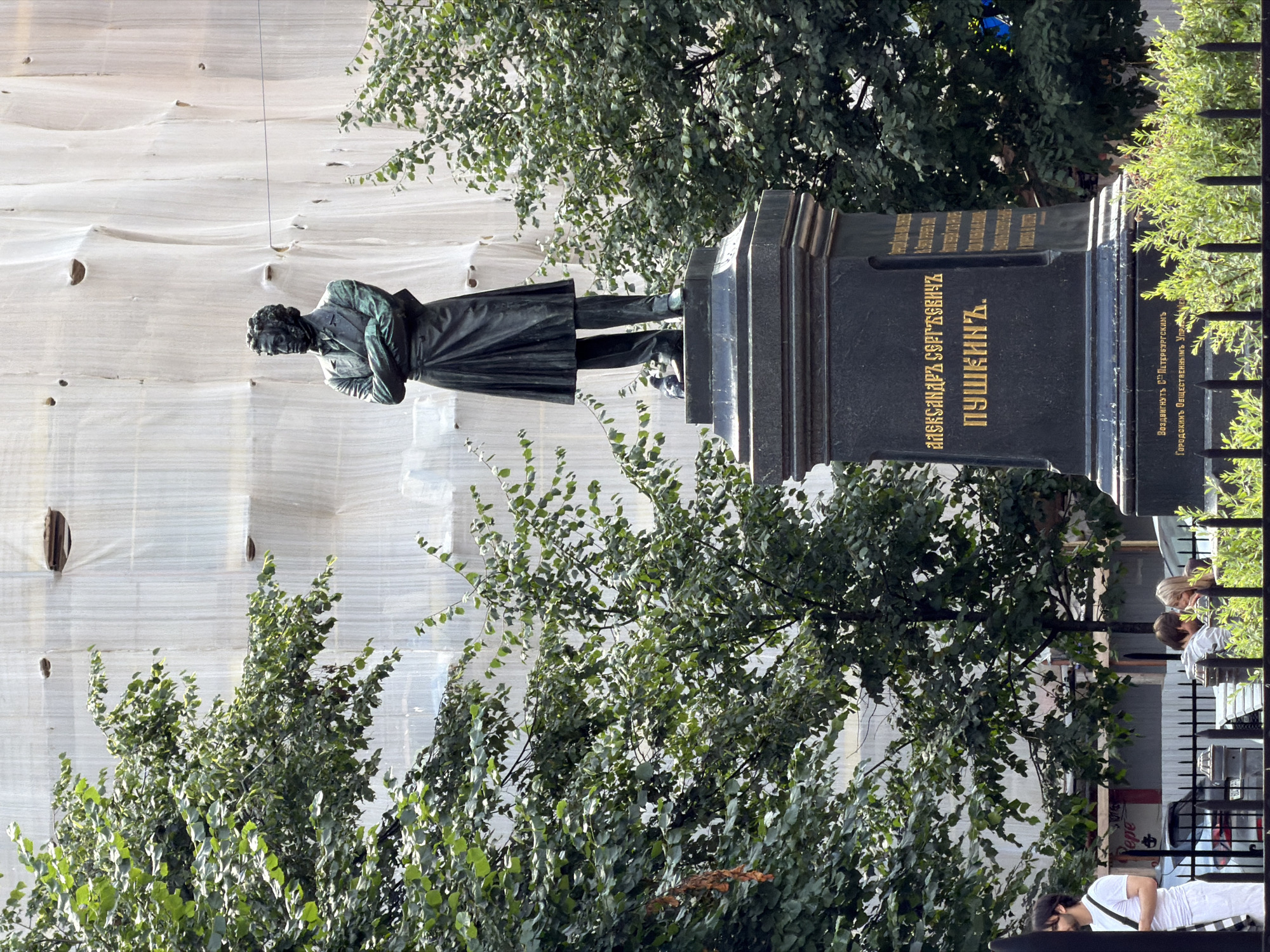 The image shows the monument to Alexander Pushkin in the city of Saint Petersburg, Russia. The monument features a bronze statue of Pushkin standing with his arms crossed, wearing a long coat and trousers. The statue is mounted on a dark granite pedestal inscribed with Russian text. The background is dominated by a large building covered in white sheeting, likely for renovation or construction. Trees with lush green foliage are visible on the left and right sides of the monument, framing the scene. In the foreground, a black metal fence separates the monument area from a pathway where a few people are visible. On the left, a man with dark hair wearing a white t-shirt and pants walks by. To the right, a couple sits at a table. Two pigeons are perched on the pedestal of the monument. The sky is not visible in the frame.