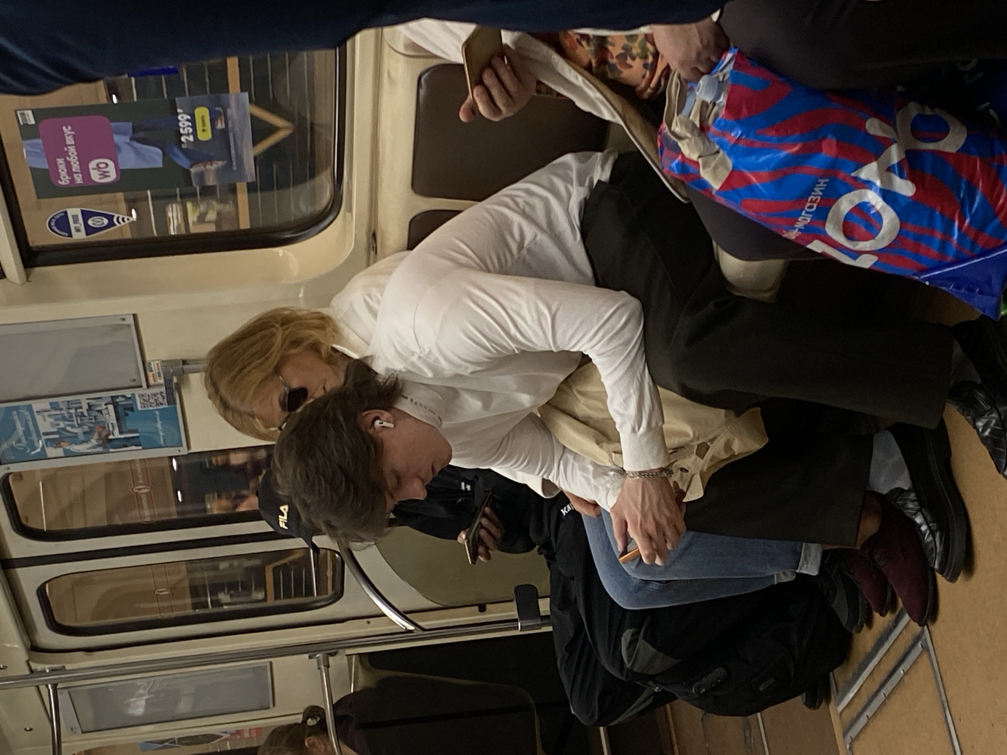 Inside a Saint Petersburg metro train, a crowded scene unfolds. A woman with blonde hair and sunglasses is seated next to a young man wearing a black FILA baseball cap and white earbuds. Both are wearing white shirts. The man is holding a phone in his hand. The man appears to be leaning on the seated woman, who seems to be resting on the seat with her eyes closed. The scene takes place on a train car. On the windows, there are advertisements and other printed posters, suggesting the interior of a public transport vehicle.
