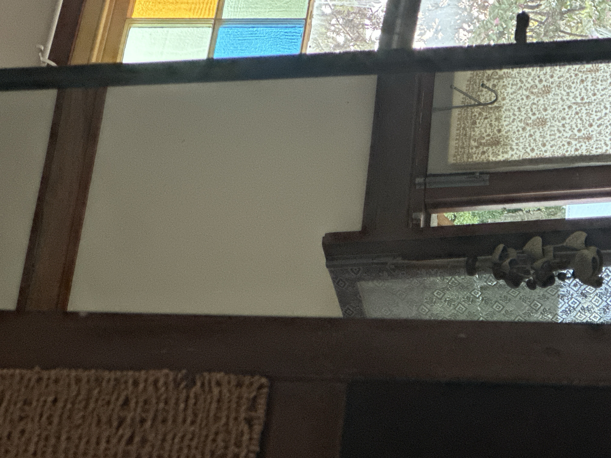 The image shows an interior view, possibly a home, with elements of a window featuring stained glass in varying colors of yellow, green, and blue. To the left, a textured woven material is visible, possibly part of a wall hanging or furniture. In the foreground, a dark, vertical wooden frame runs from top to bottom, obscuring a portion of the scene. Further into the image, a patterned screen or curtain is partially visible, with a hanging object resembling a wind chime or decorative item. The background reveals more of the stained glass window, suggesting it is a significant architectural feature. The environment appears to be well-lit, with natural light filtering through the window. There is no indication of people or specific actions taking place. The location in Thora, Australia, is not discernible from the visual information provided in the image.