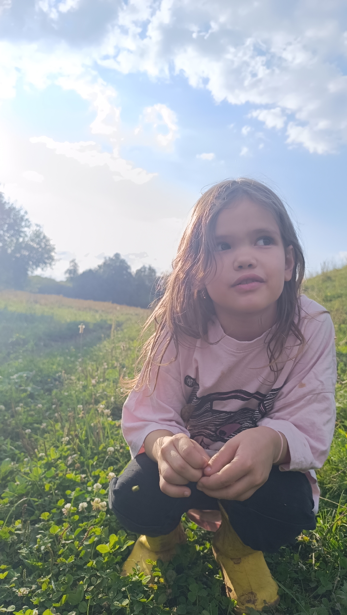 A young girl with long, brown hair is crouching in a grassy field. She is wearing a pink long-sleeved shirt, black pants, and yellow boots. The girl is looking to her right, with her hands clasped in front of her. The background shows a gentle slope of green grass, with trees and a cloudy blue sky. The overall lighting suggests it is daytime, possibly late afternoon, with a slight sun flare visible. The environment appears to be an outdoor, natural setting. There is no discernible urban infrastructure visible to indicate a specific location within the city of Karsha, Russia.