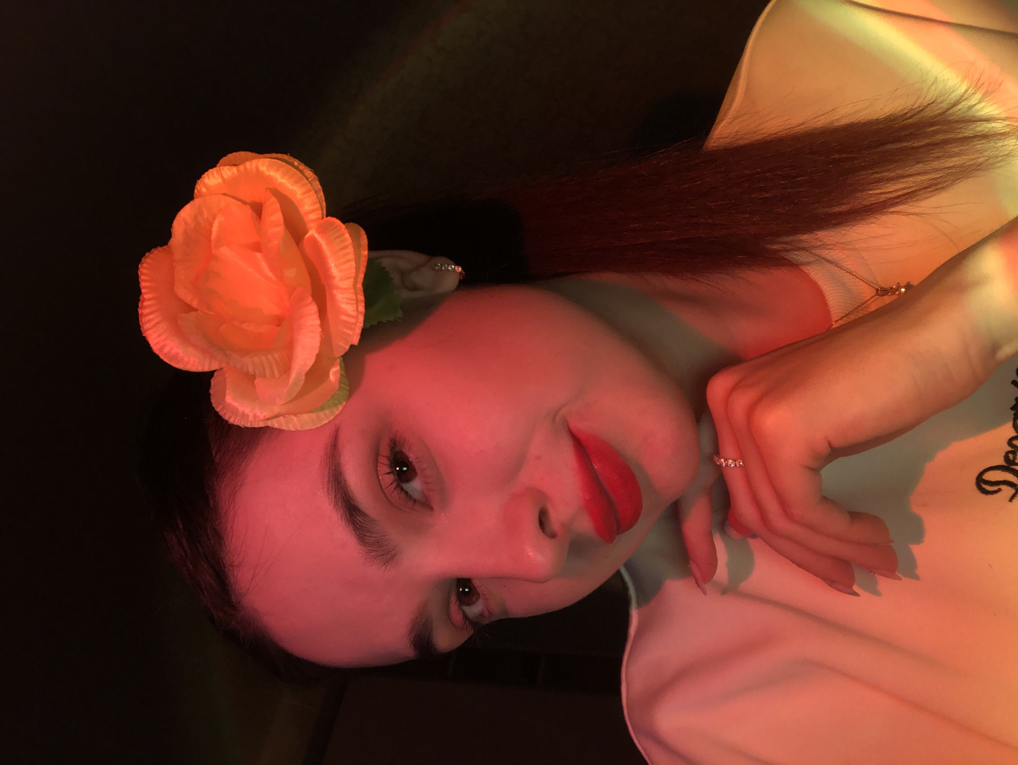 A young woman with shoulder-length brown hair is posing with her head tilted. She is wearing a light-colored shirt. A large orange rose is positioned near her ear. She is wearing red lipstick and what appears to be a diamond ring. The background is dark. The location is not discernible from the provided image.